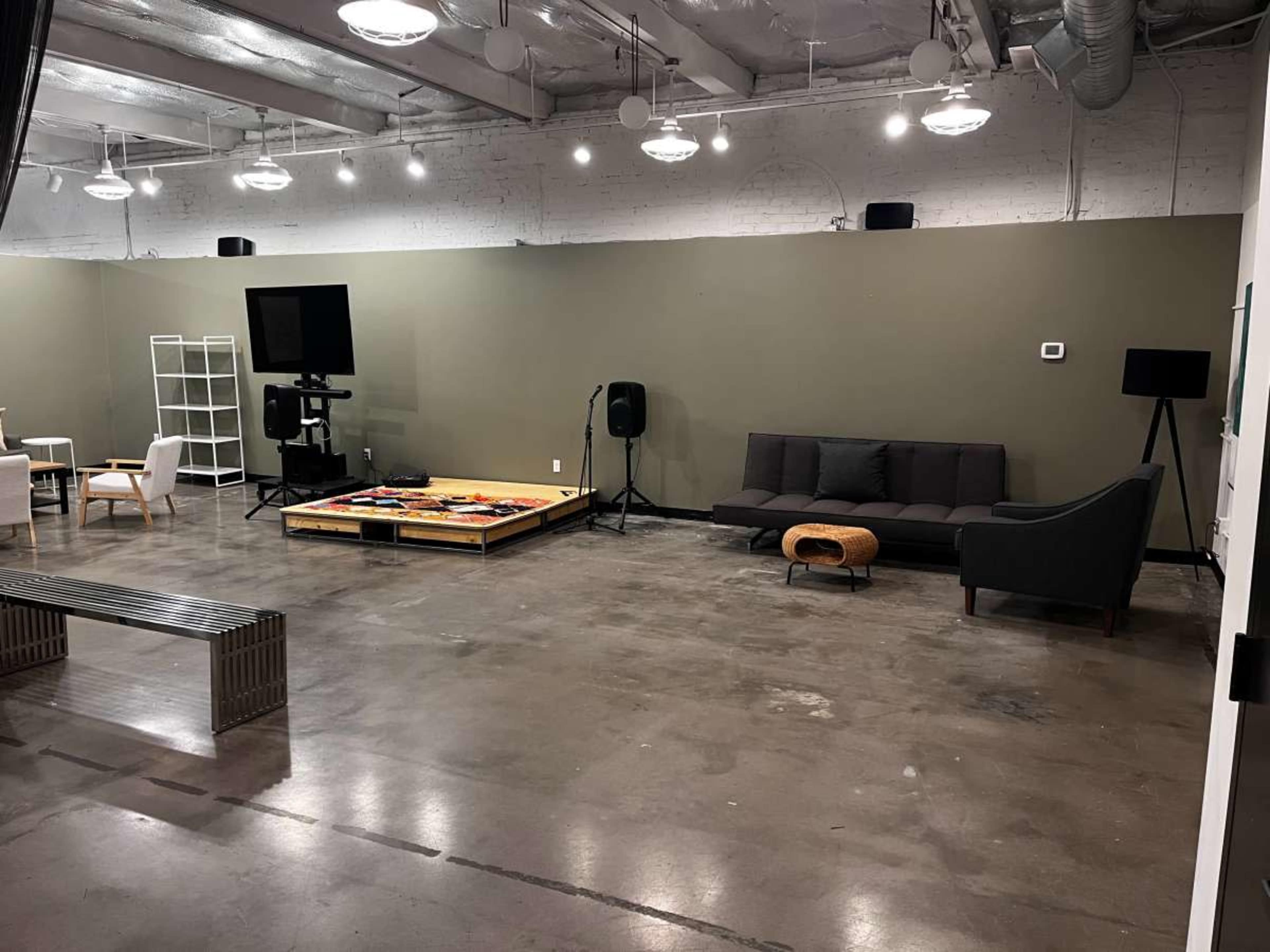 The image shows a spacious, minimalist room with concrete flooring, a gray wall, various seating areas, and a coffee table.