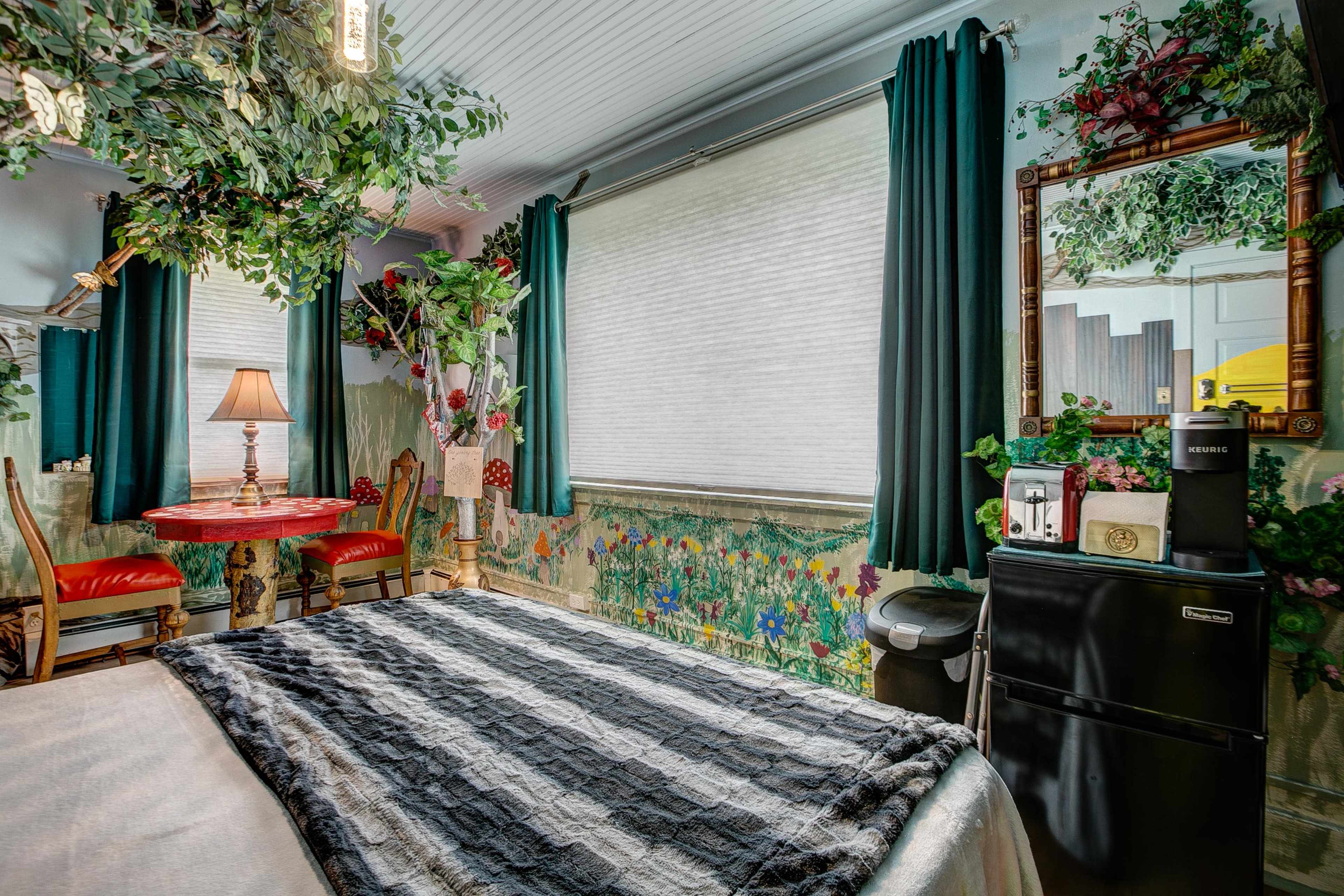 A cozy bedroom featuring a bed with a striped blanket, a small dining table with red chairs, a decorative mirror, and a mini-fridge, all surrounded by floral wallpaper and indoor plants.