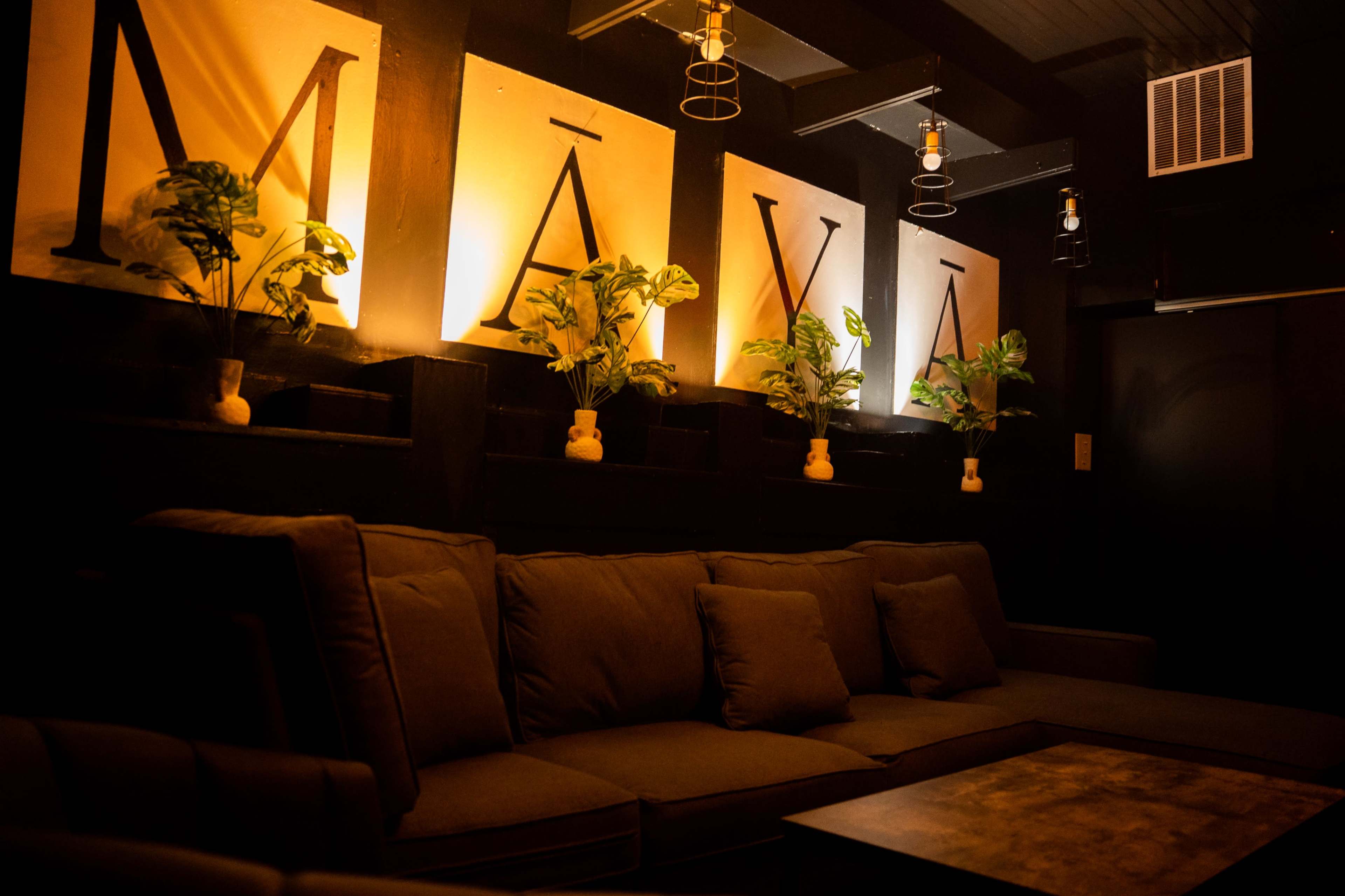 The image shows a lounge area with dark walls, featuring large illuminated signs that spell "MĀYĀ" and potted plants beside them.