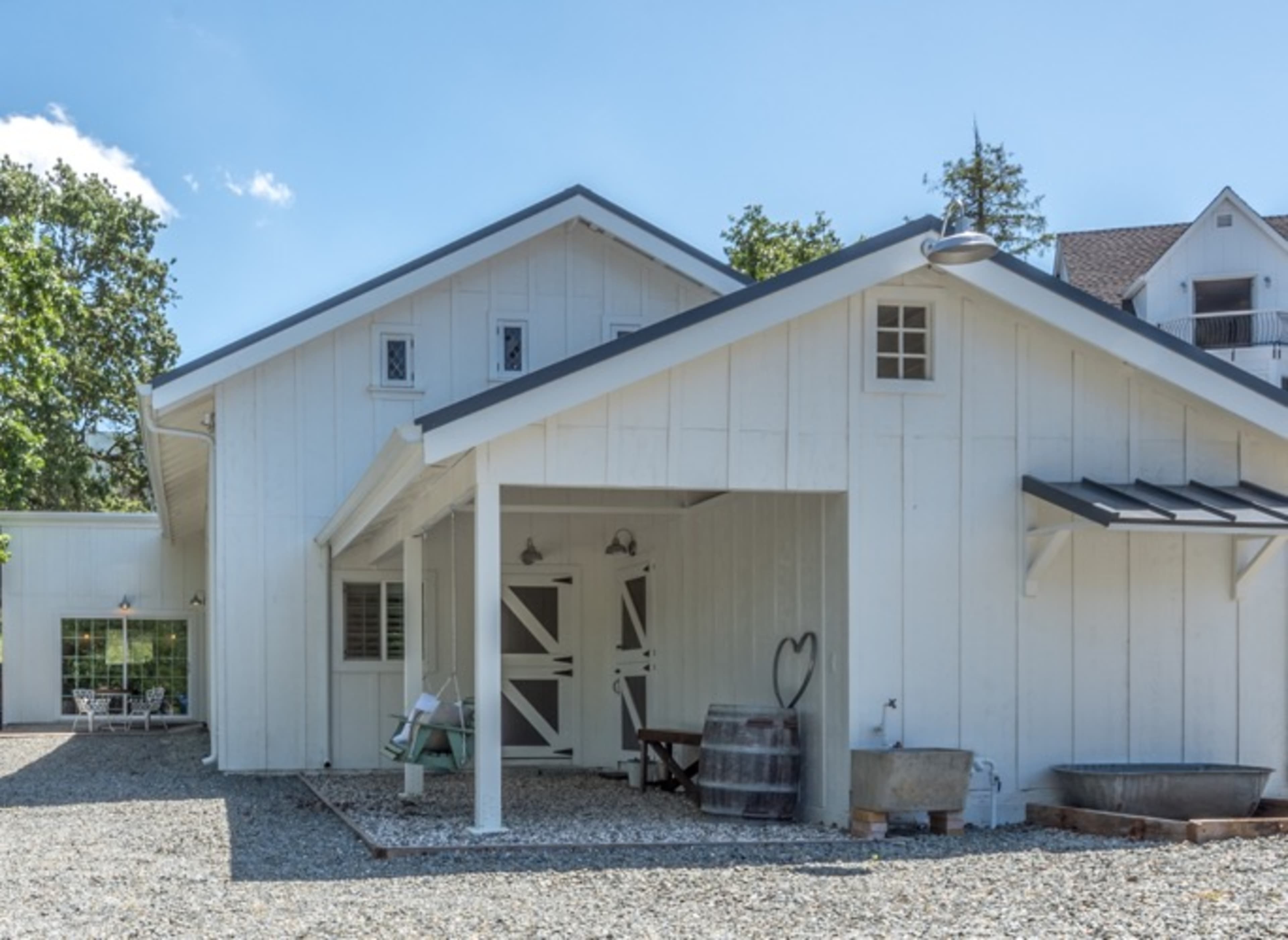A white, wooden farmhouse with a sloped roof, surrounded by gravel and trees.