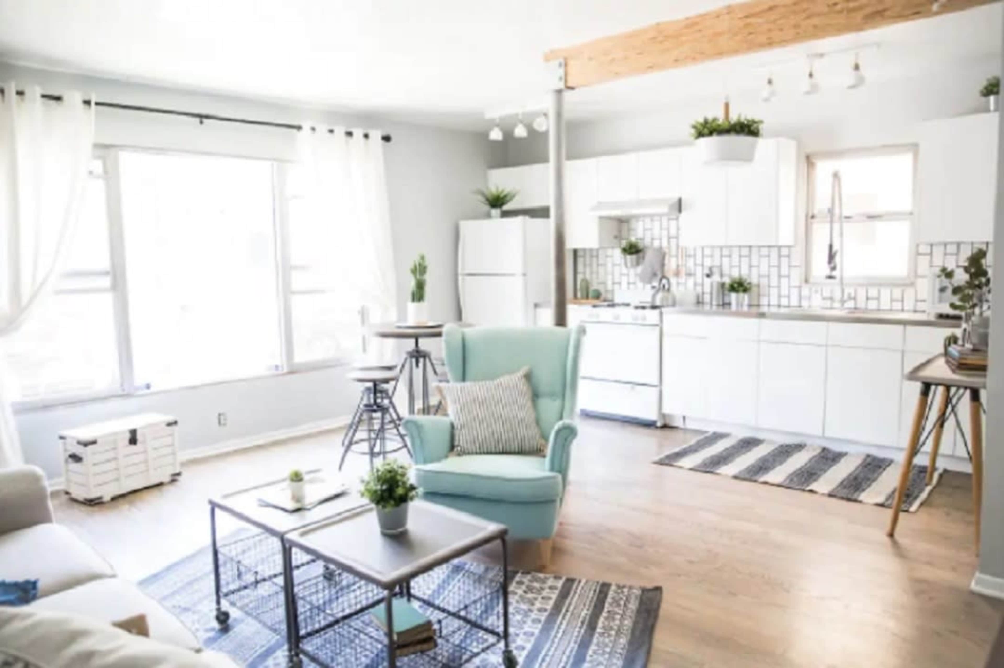 The image shows a modern, open-concept living space featuring a light-colored kitchen, a cozy seating area with a teal armchair, and natural light streaming through large windows.