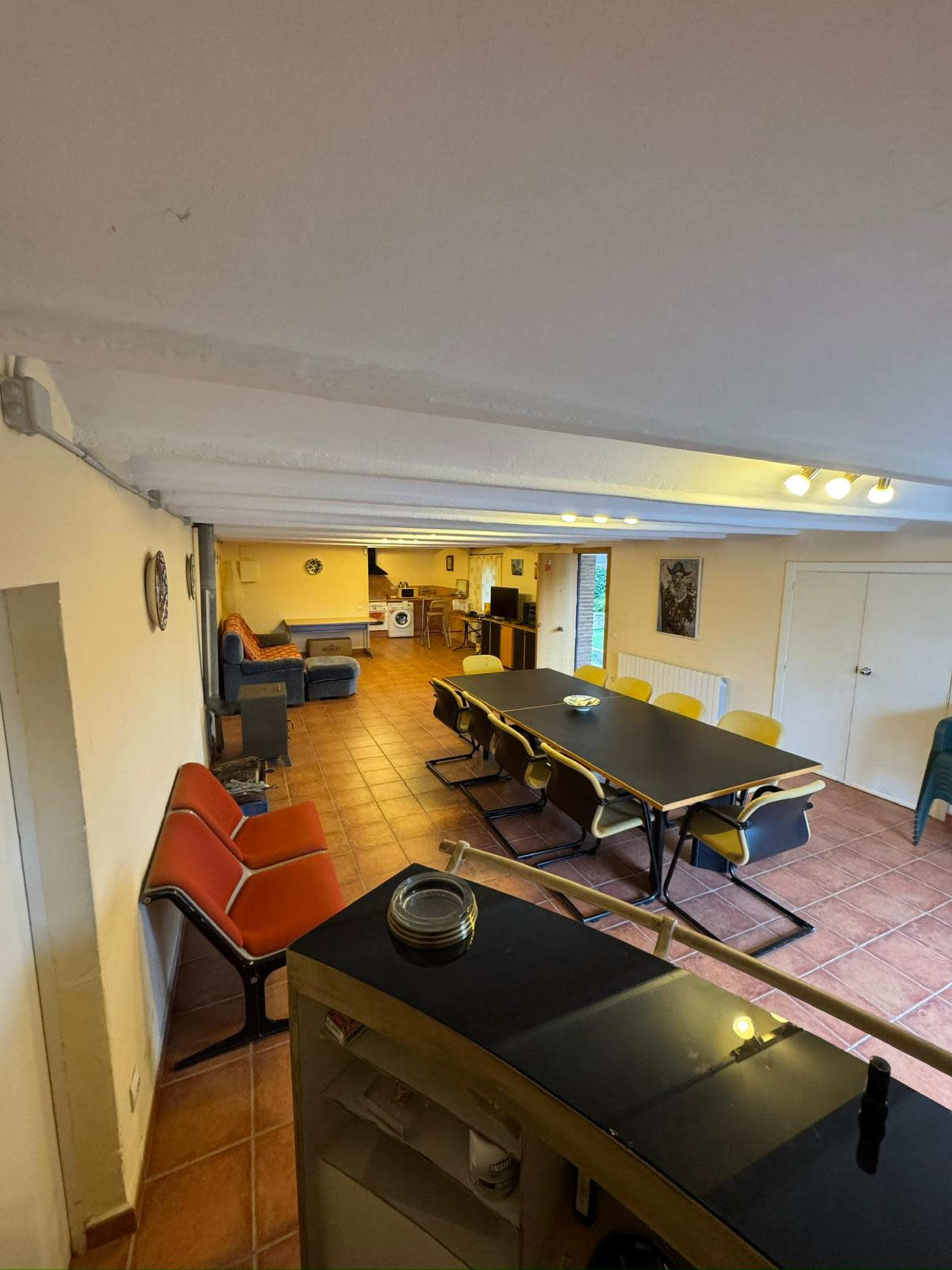The image shows a spacious room with a long black table surrounded by chairs, a seating area with sofas, and an open kitchen in the background.