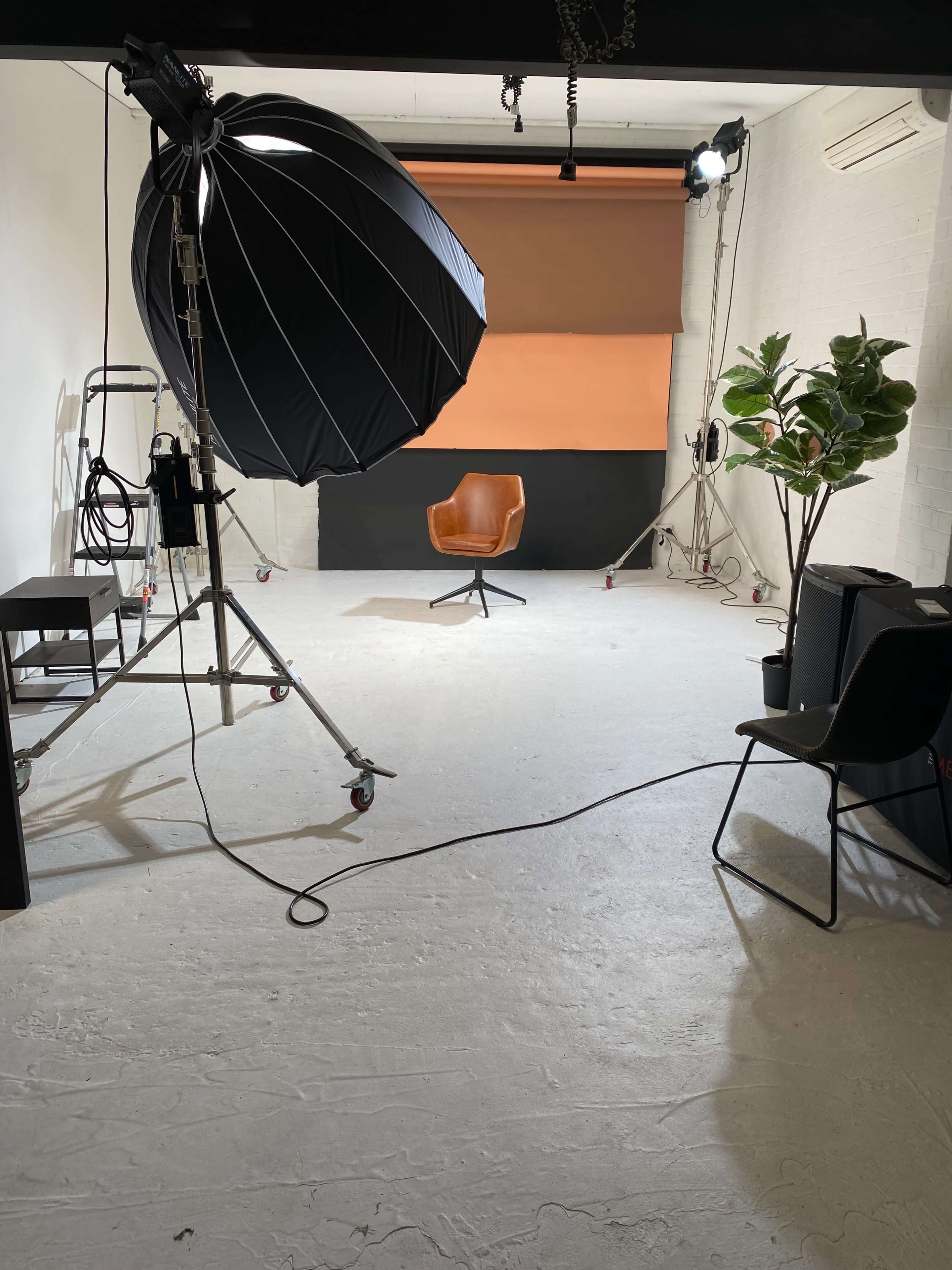 A photography studio features a large umbrella light, an empty orange-backed set, a brown chair, and various equipment.
