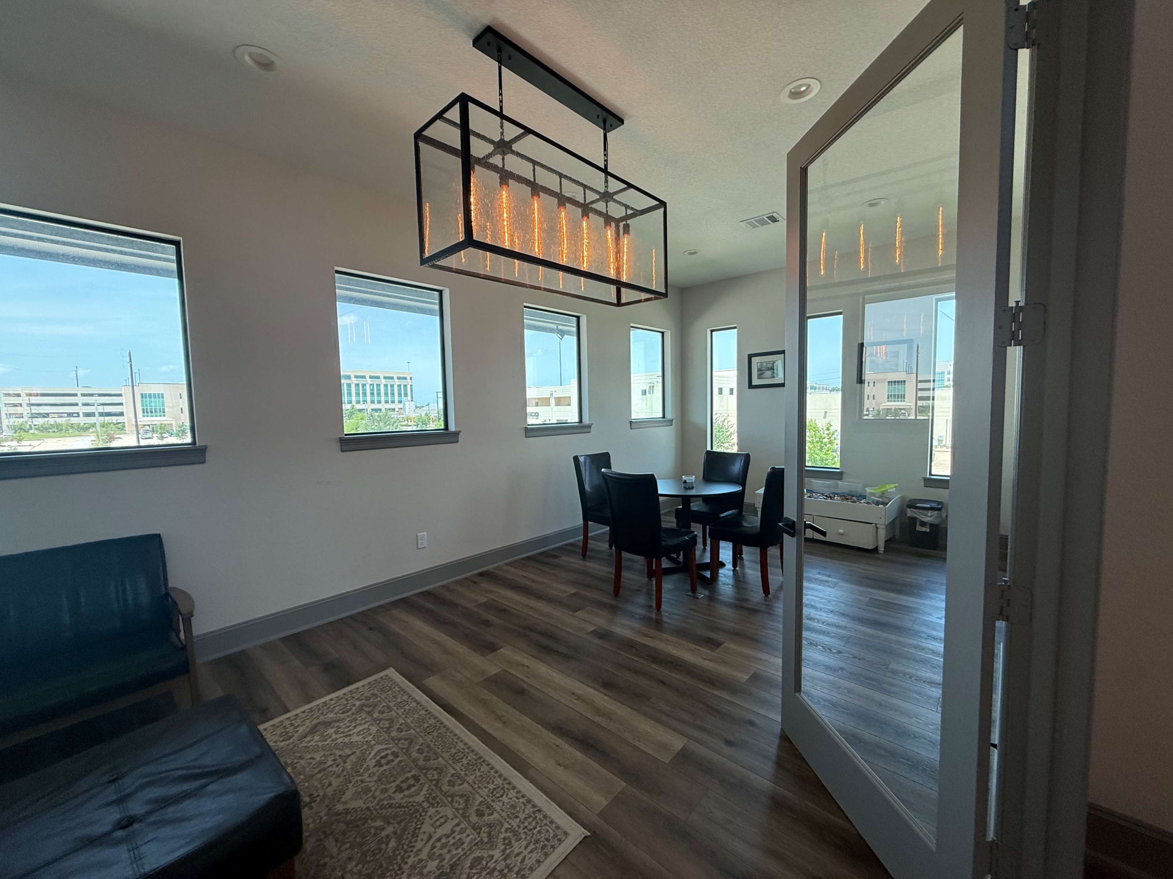 The image shows a modern, well-lit room with a dining table and chairs, large windows, and a stylish light fixture.