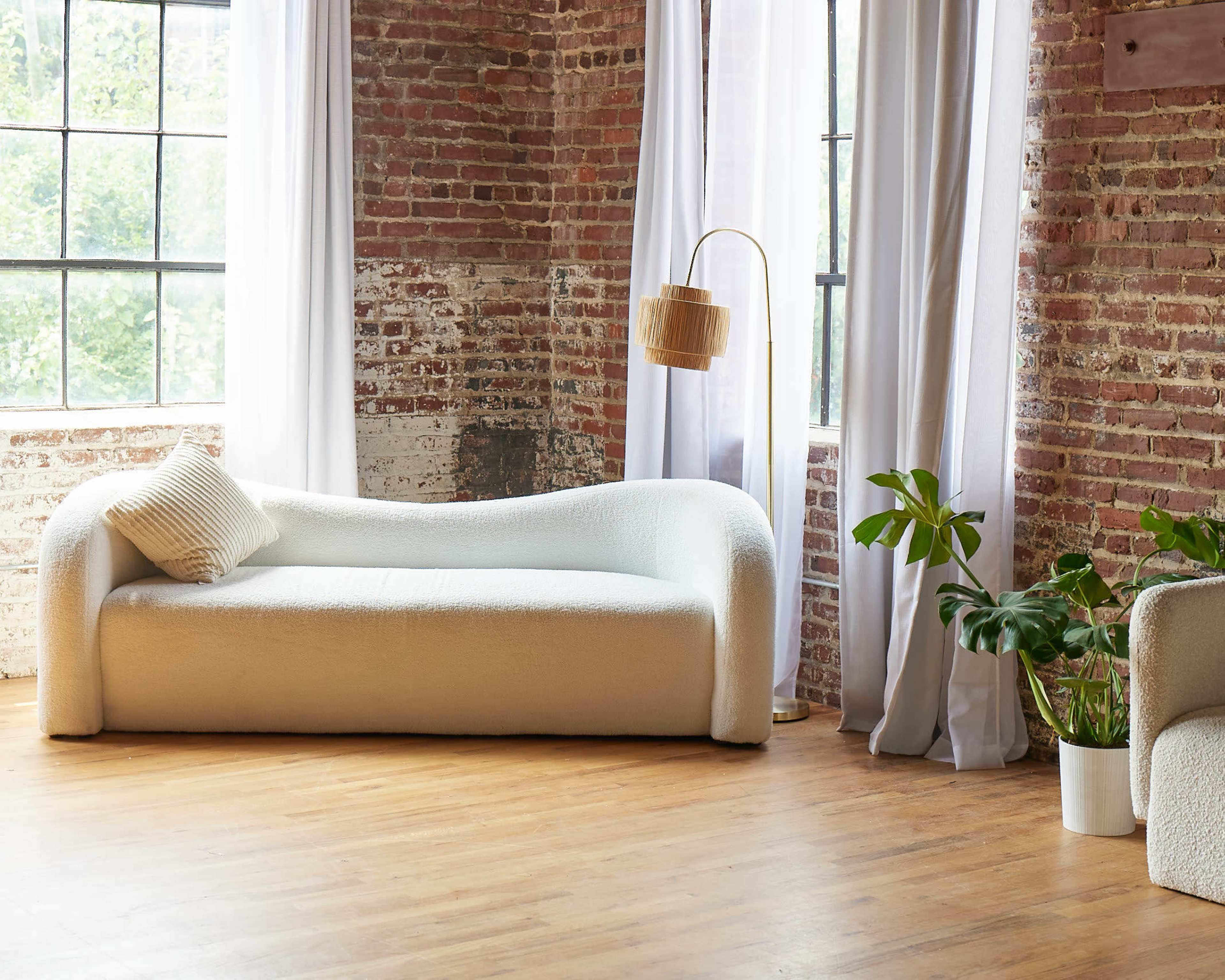 A white curved sofa is placed in a room with exposed brick walls and large windows, accompanied by a tall floor lamp and a potted plant.