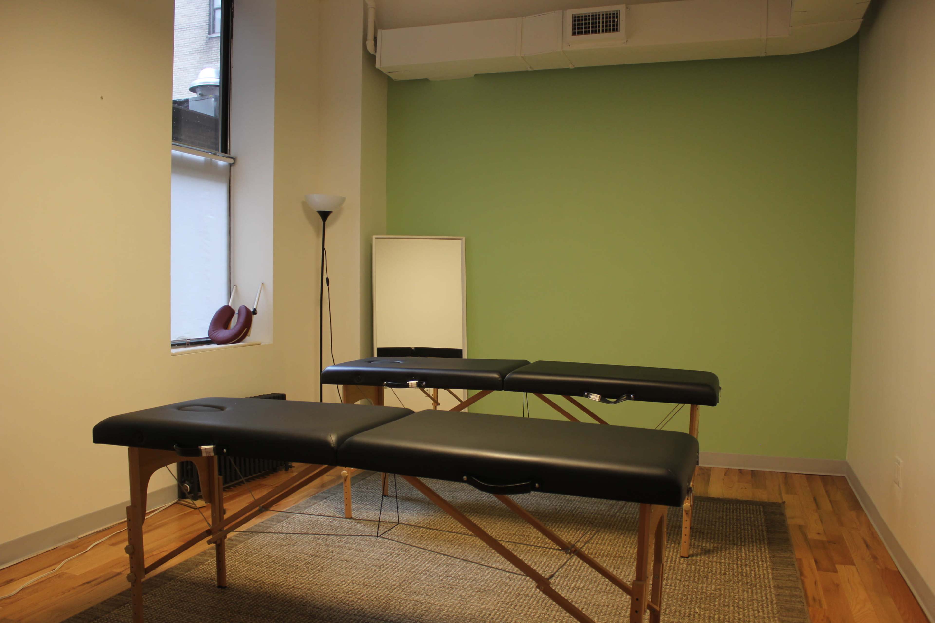 A sparsely furnished therapy room featuring two black massage tables on a natural fiber rug against a green wall.