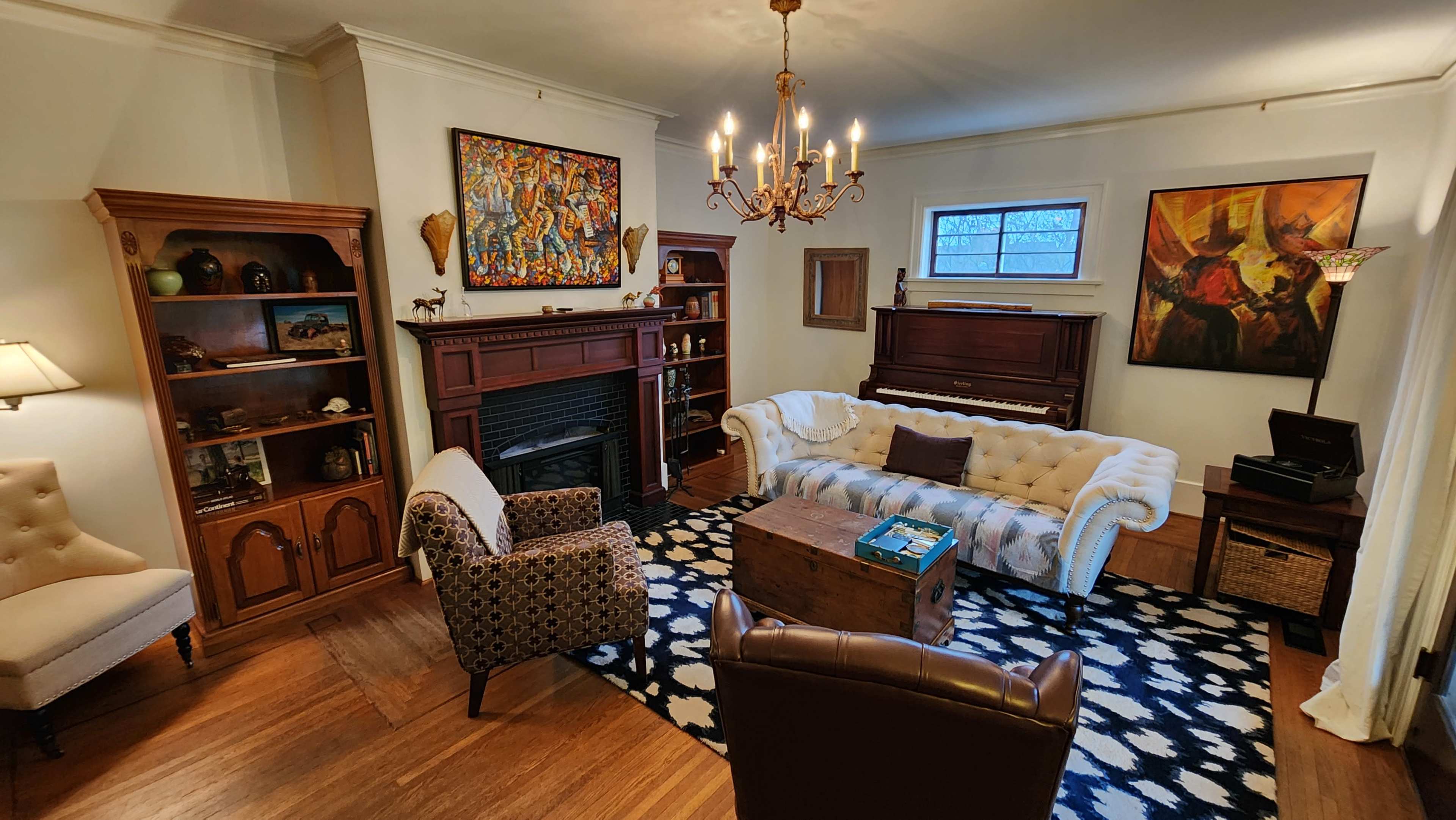 A cozy living room features a white tufted sofa, a wooden coffee table, and two armchairs, surrounded by bookshelves and art on the walls.