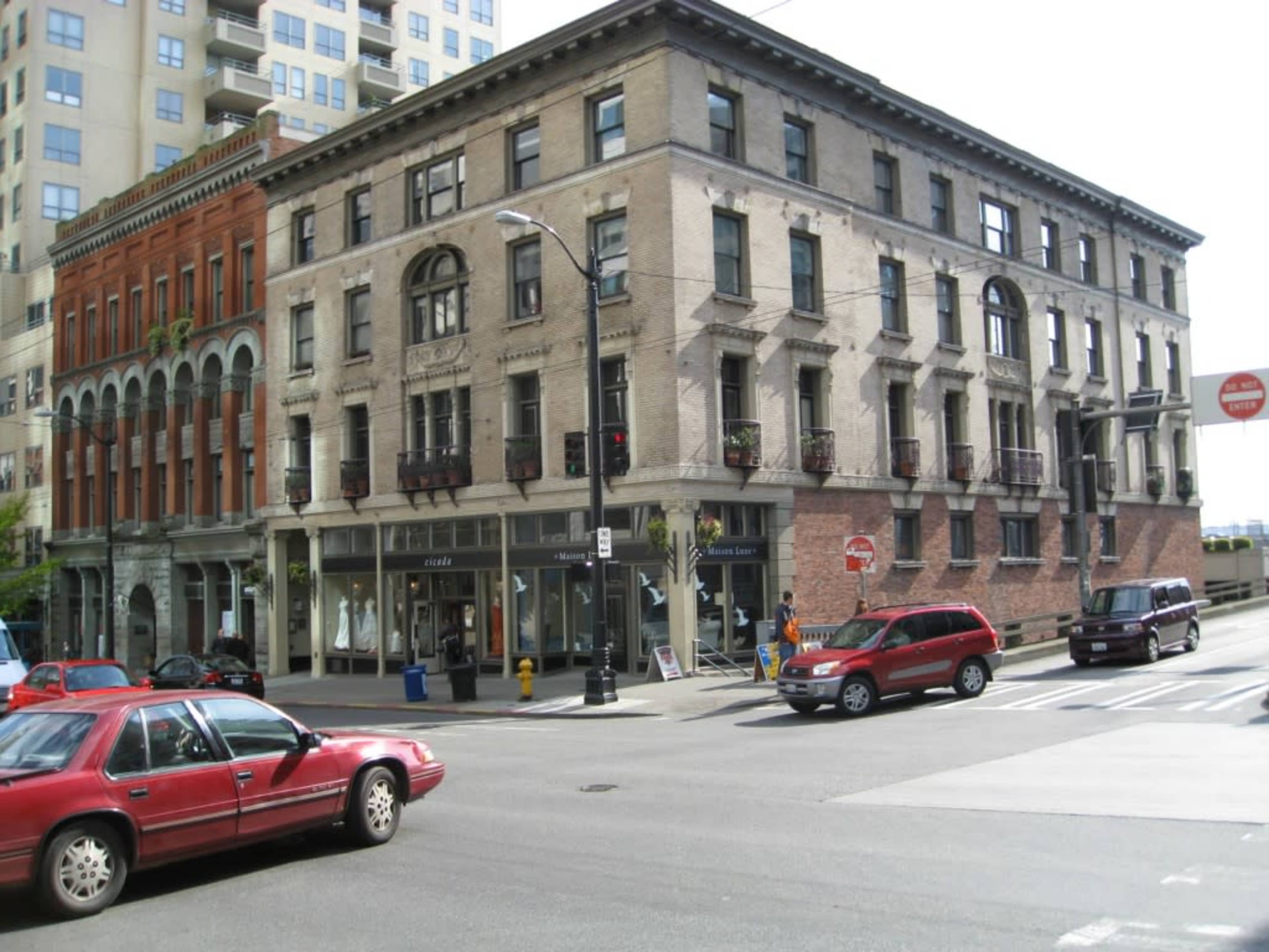 The image shows a street corner with a multi-story brick building featuring large windows and balconies, adjacent to a taller modern building, with cars parked and driving along the road.