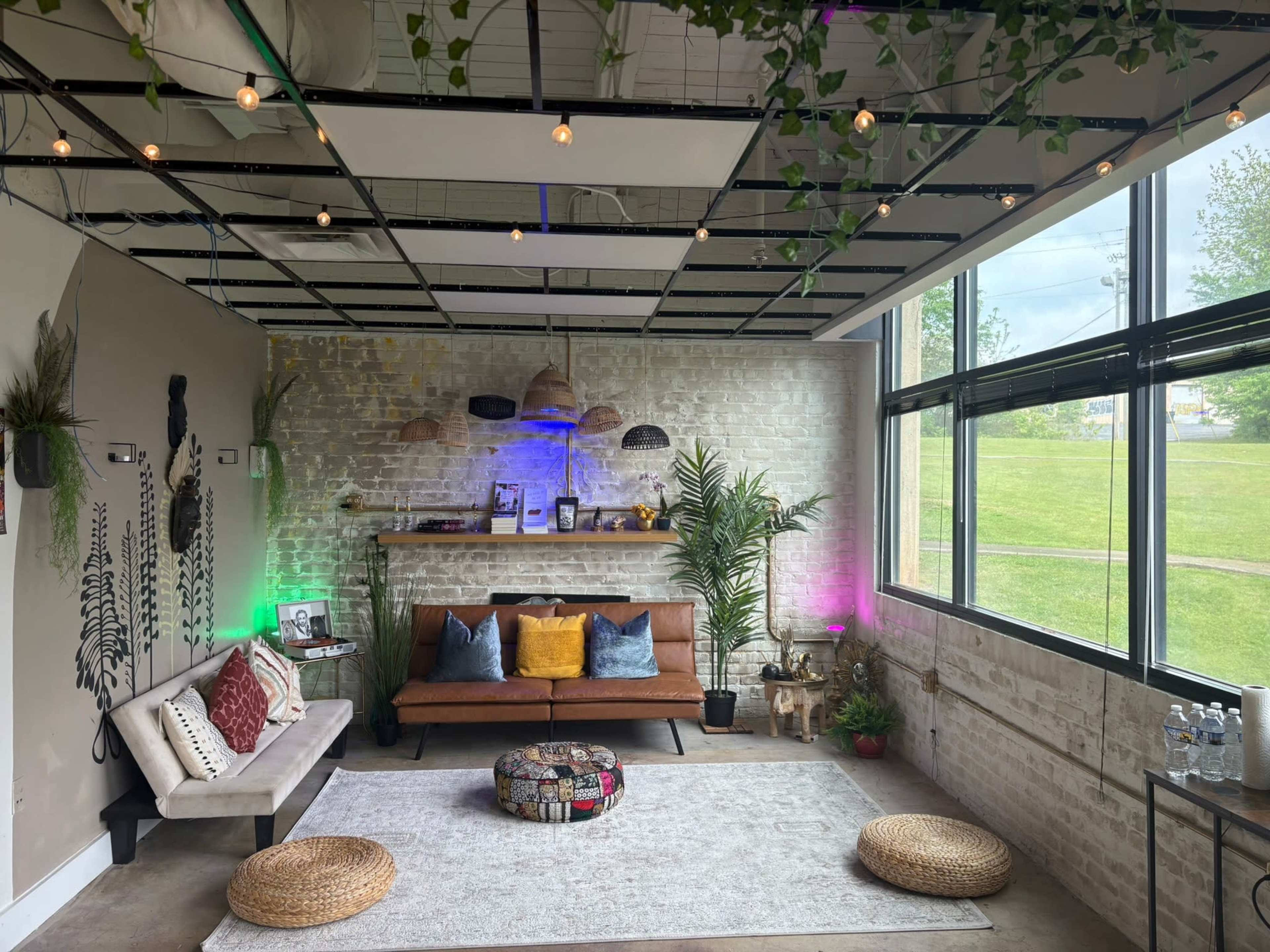 The image shows a cozy indoor lounge area featuring a brown sofa, a decorative rug, a fireplace shelf, and plants, illuminated by colored lights.