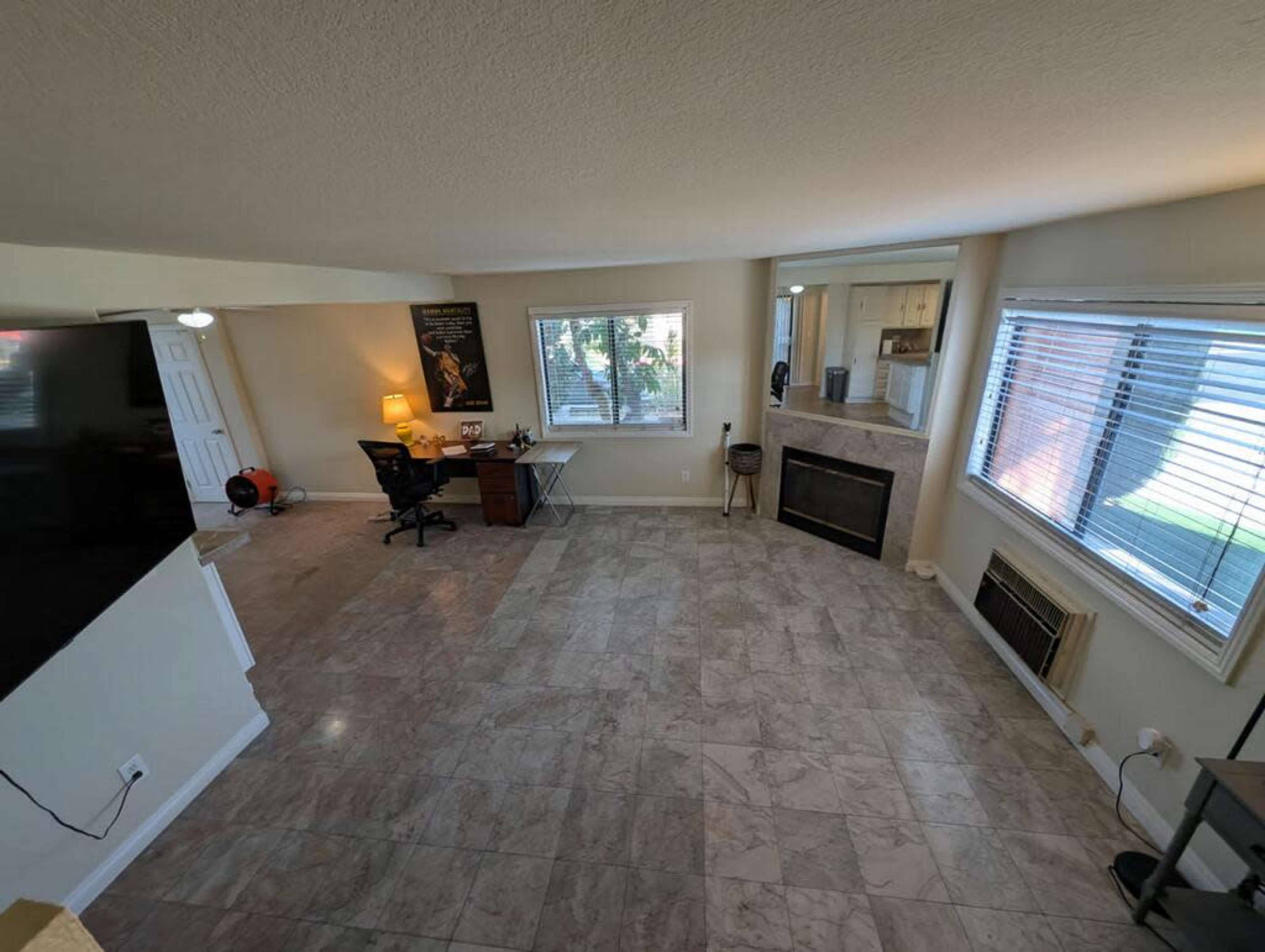 The image shows a spacious living room with tiled floors, a desk and chair, a fireplace, and large windows allowing natural light to enter.