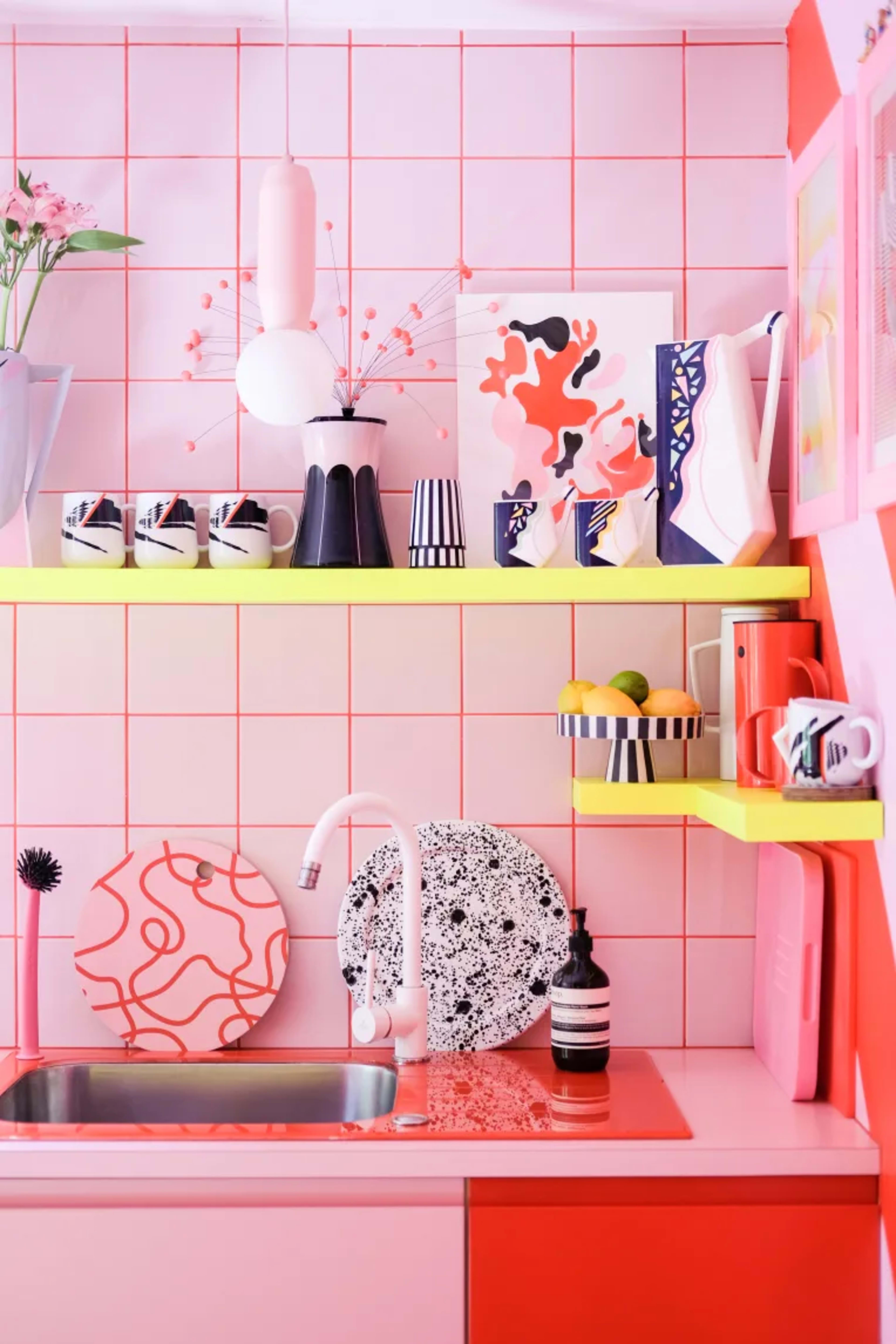 A vibrant pink kitchen features patterned dishware and art displayed on colorful shelves above a sink.