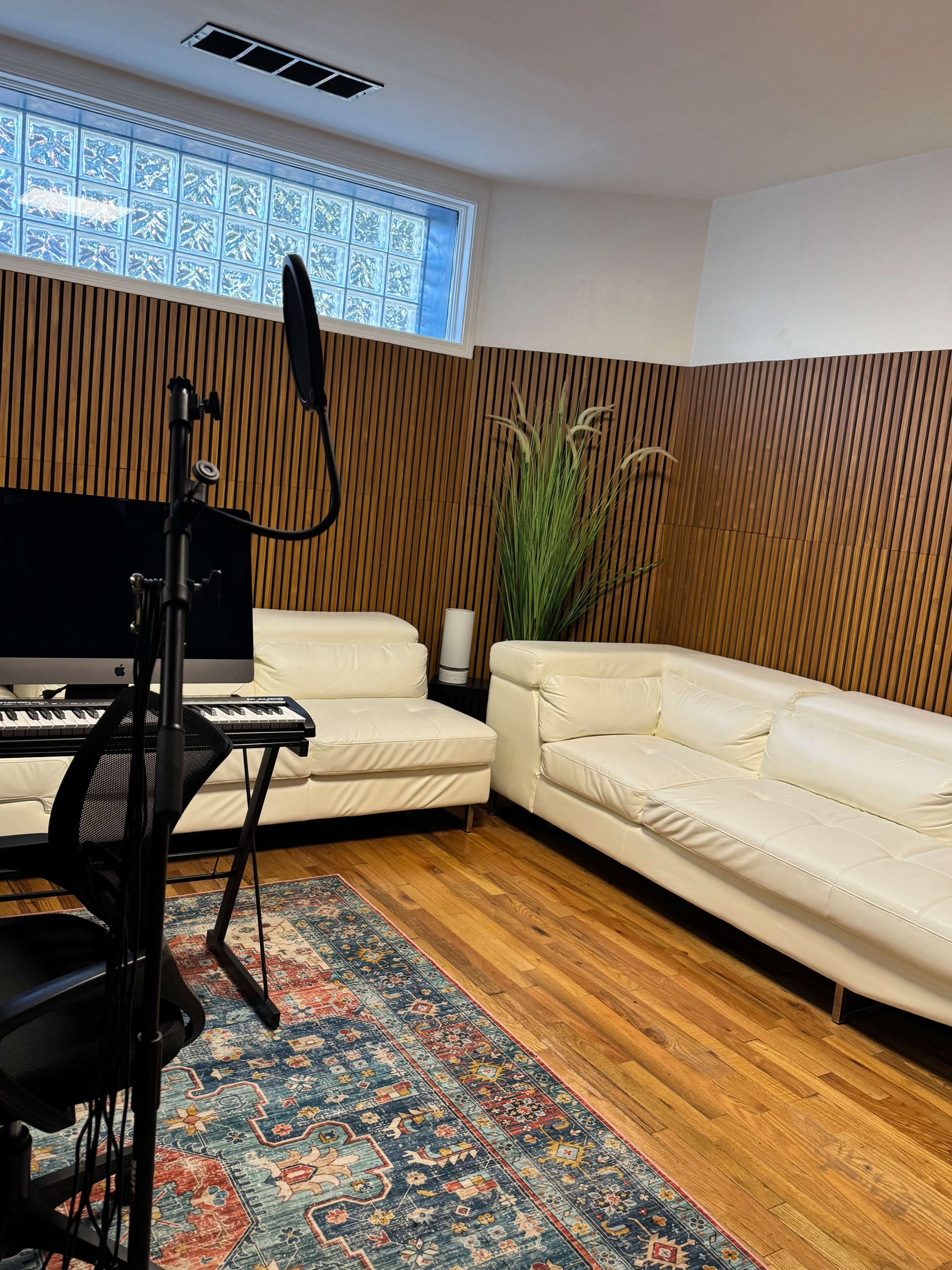 The image shows a cozy recording studio featuring two white leather sofas, a microphone on a stand, a keyboard, and a colorful area rug on wooden flooring.