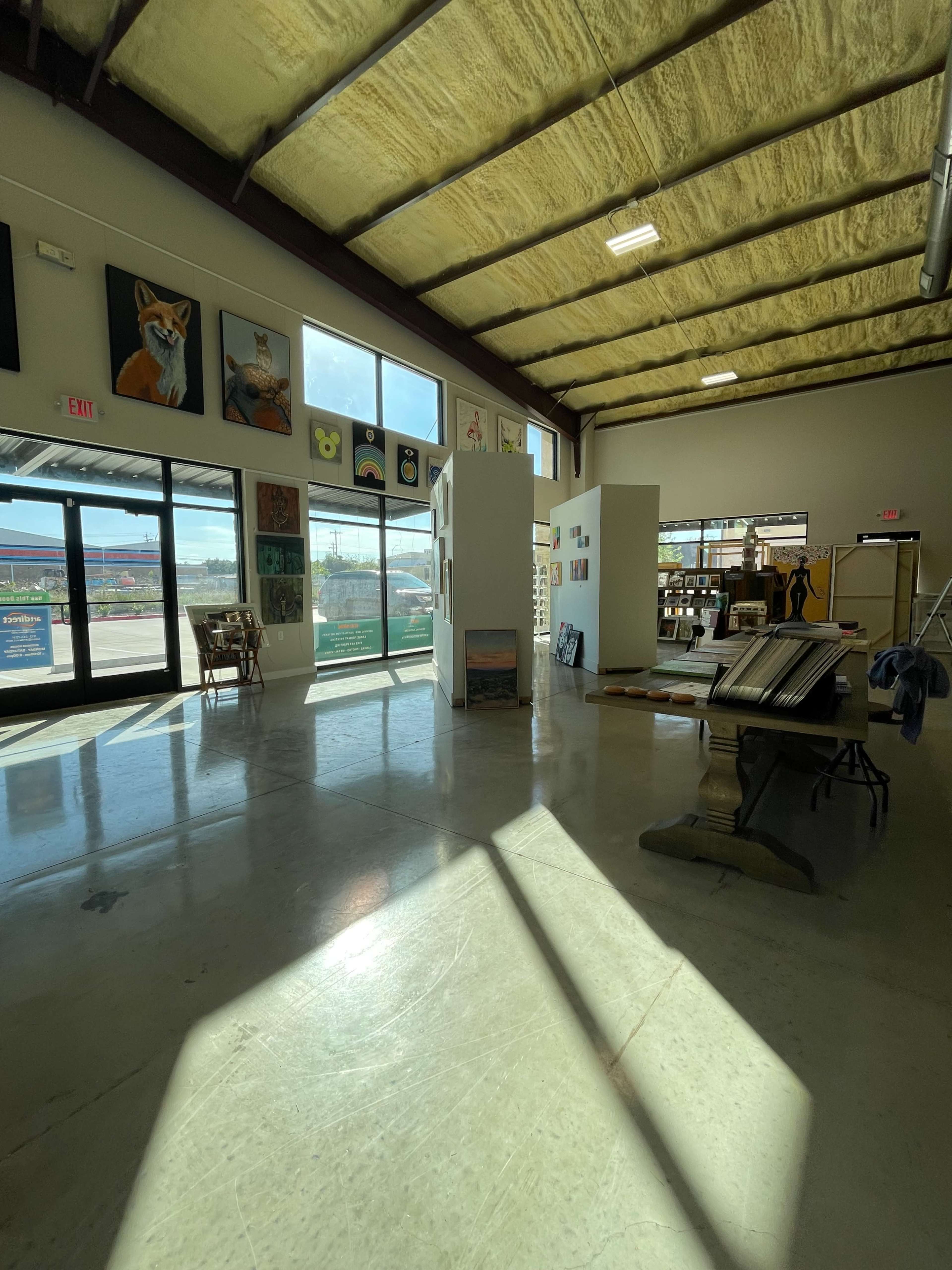 The interior of a spacious art gallery features white walls adorned with various artworks and large windows allowing natural light to illuminate the space.