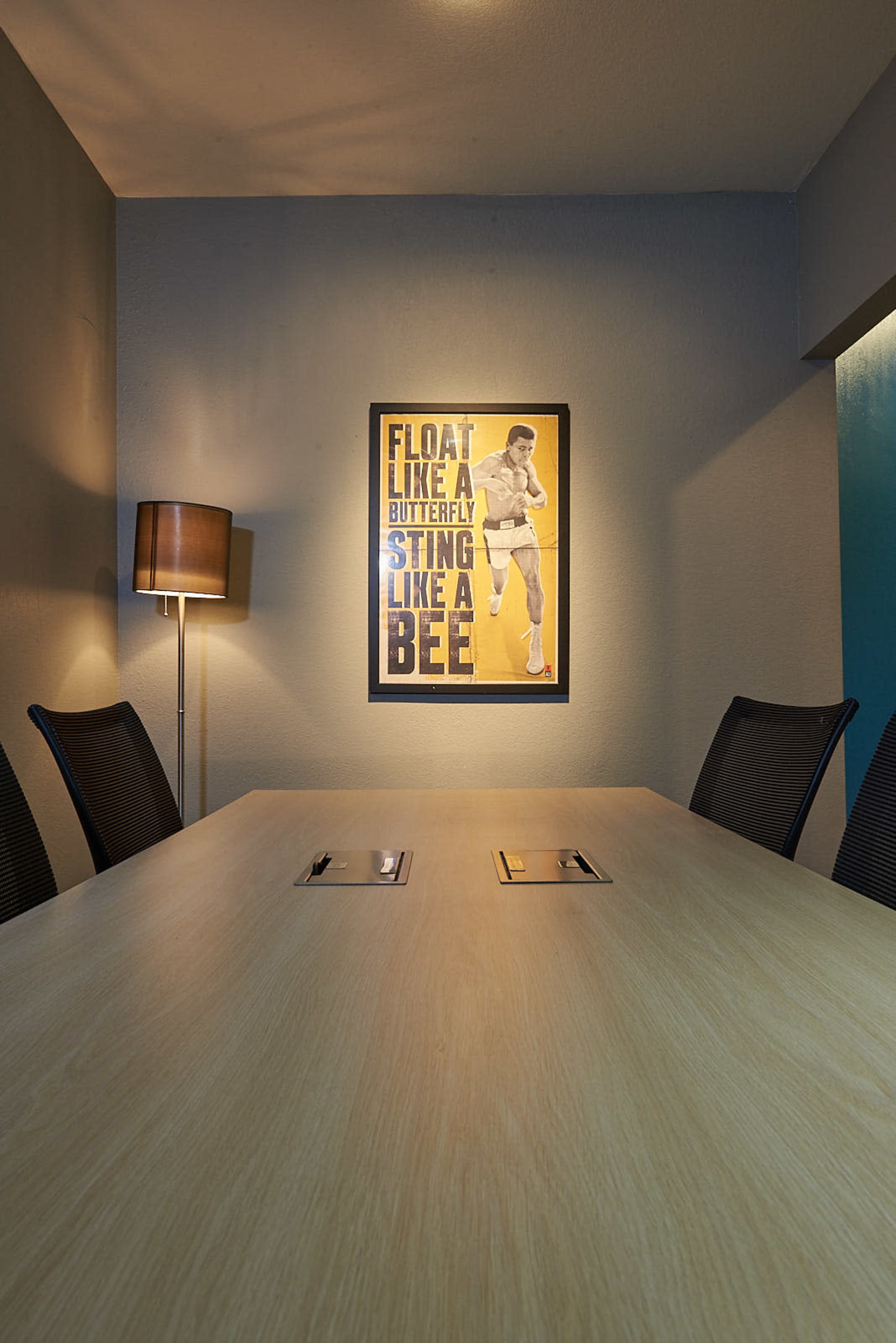 A meeting room features a wooden table surrounded by chairs, with a lamp and a motivational poster on the wall.