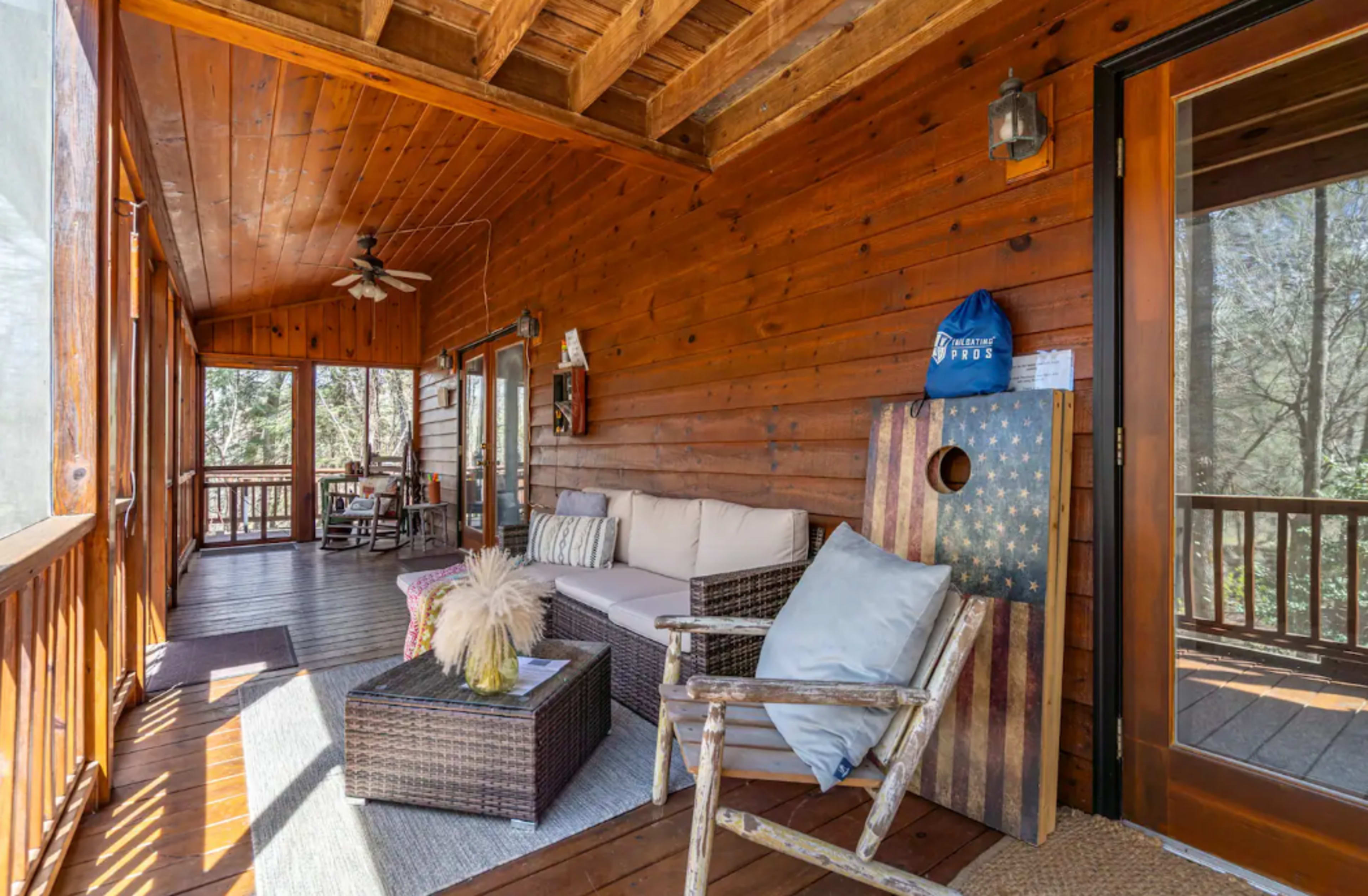 A wooden porch with a seating area that includes a couch, a coffee table, and an armchair, surrounded by large windows and natural light.