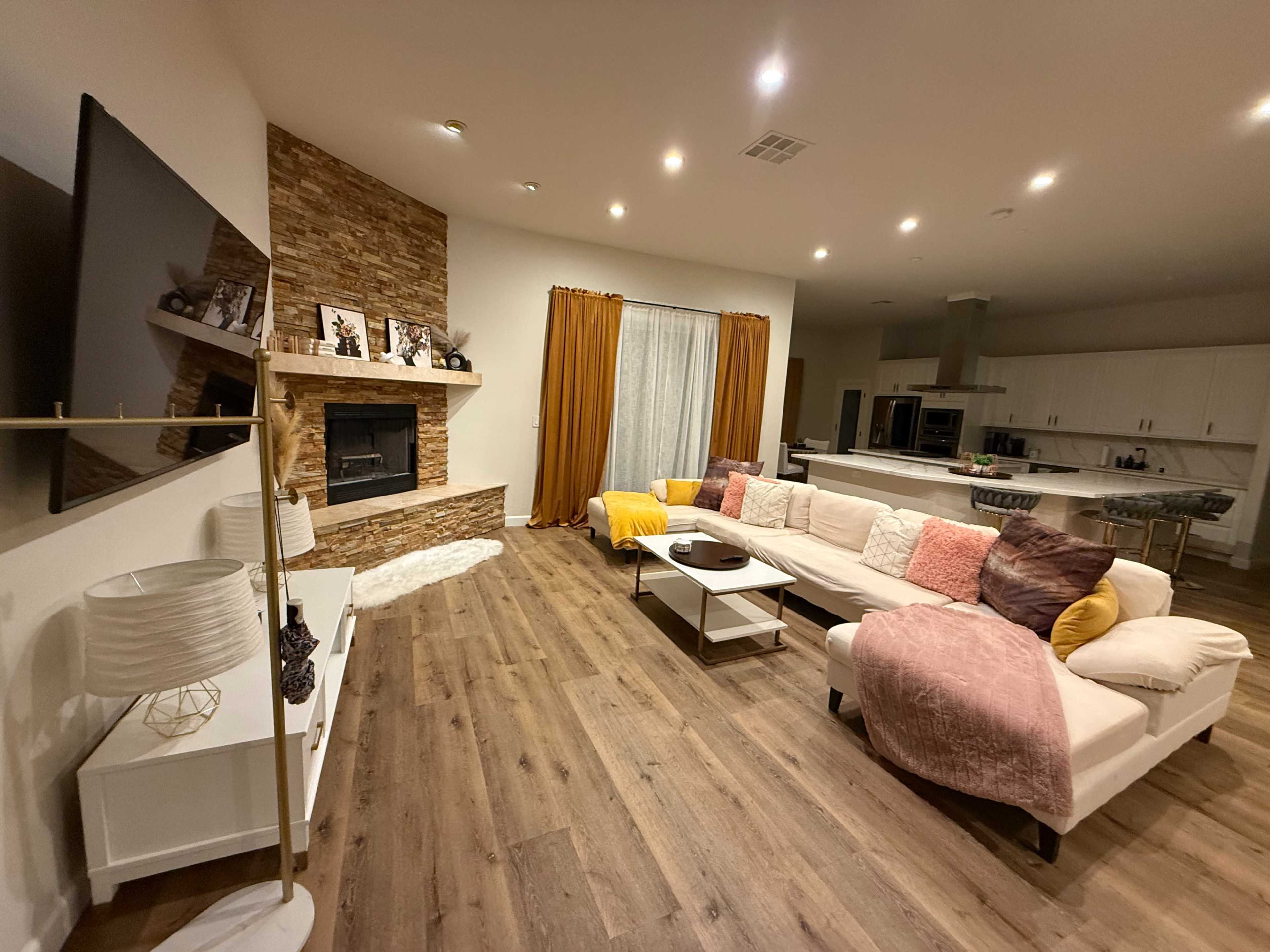 The image shows a modern living room with a large sectional sofa, a stone fireplace, and kitchen visible in the background, featuring white cabinetry and warm lighting.