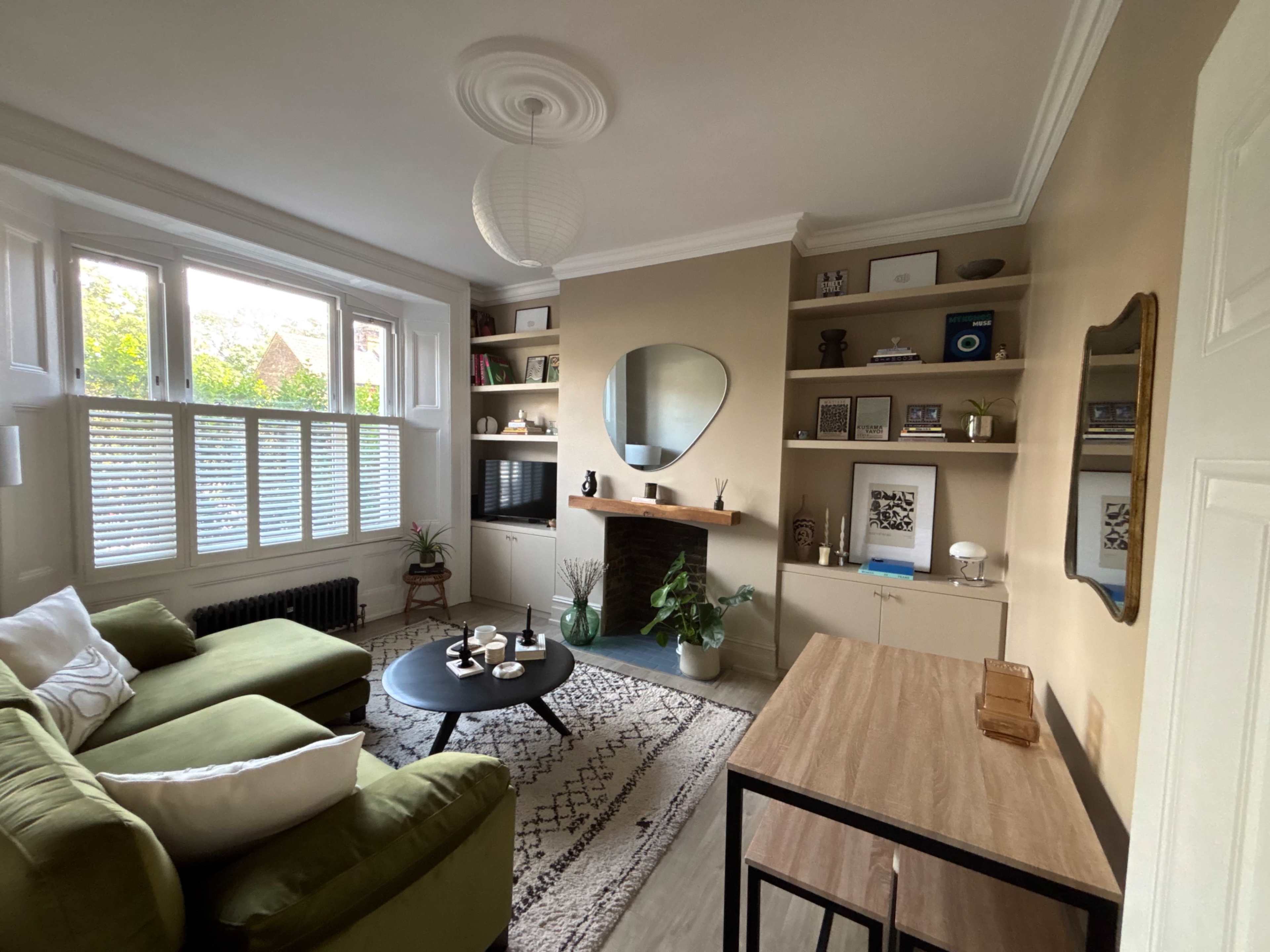 The image shows a cozy living room with a green sectional sofa, a round black coffee table, a fireplace, and a wall of shelves displaying various decorative items.