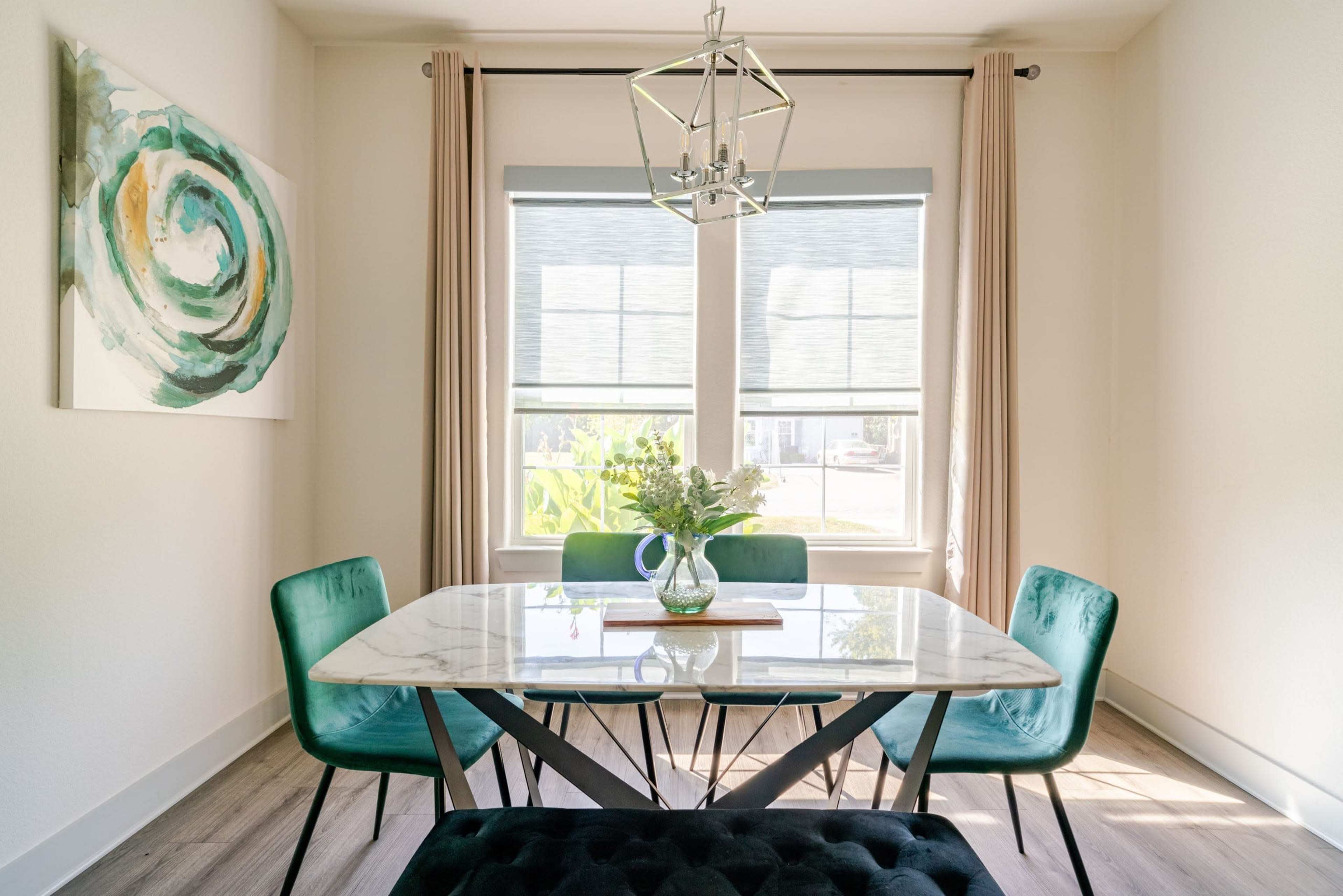 A modern dining area features a marble-top table surrounded by green chairs, with large windows and a decorative light fixture above.