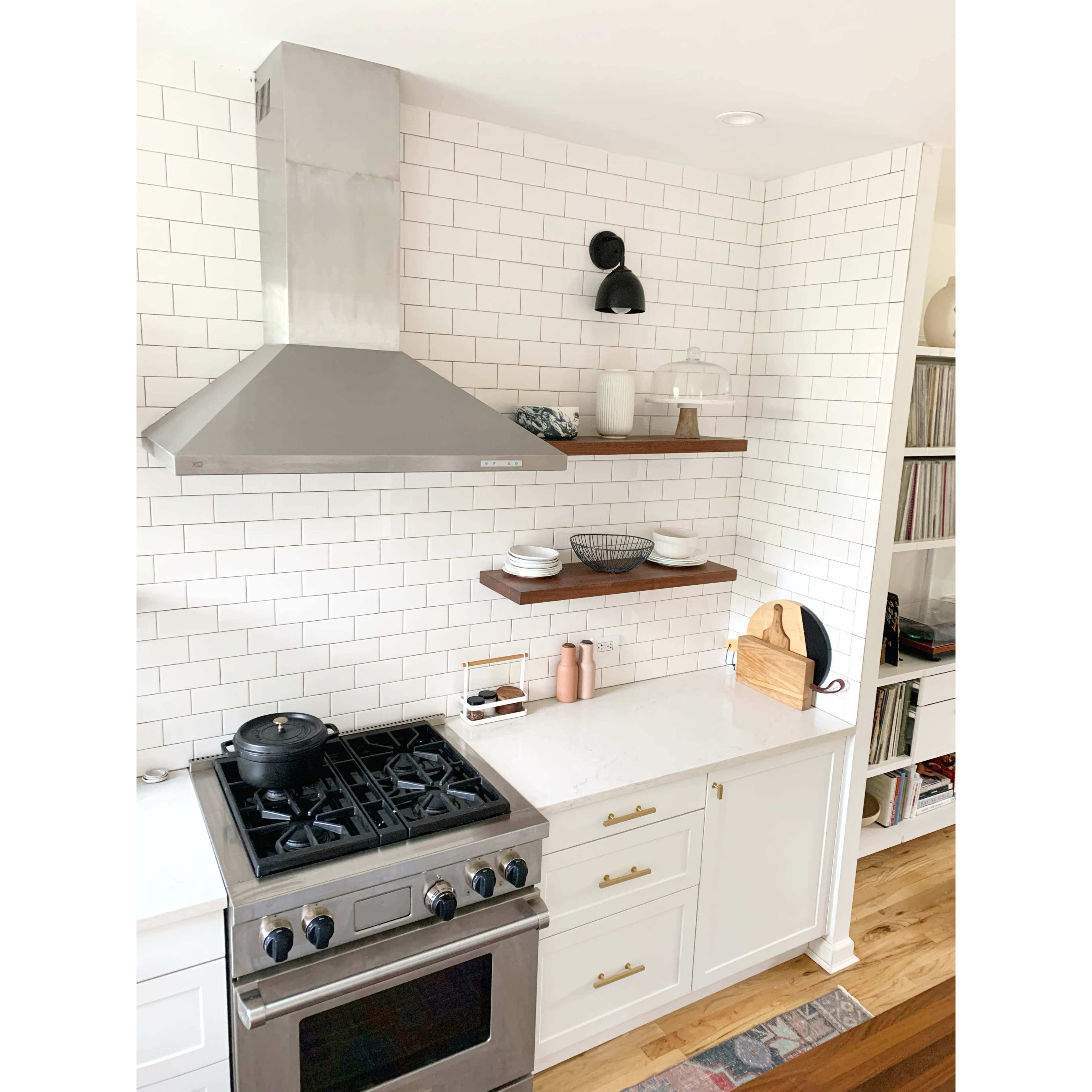A modern kitchen with a stainless steel range hood, white subway tile backsplash, wooden open shelves, and a gas stove beneath.