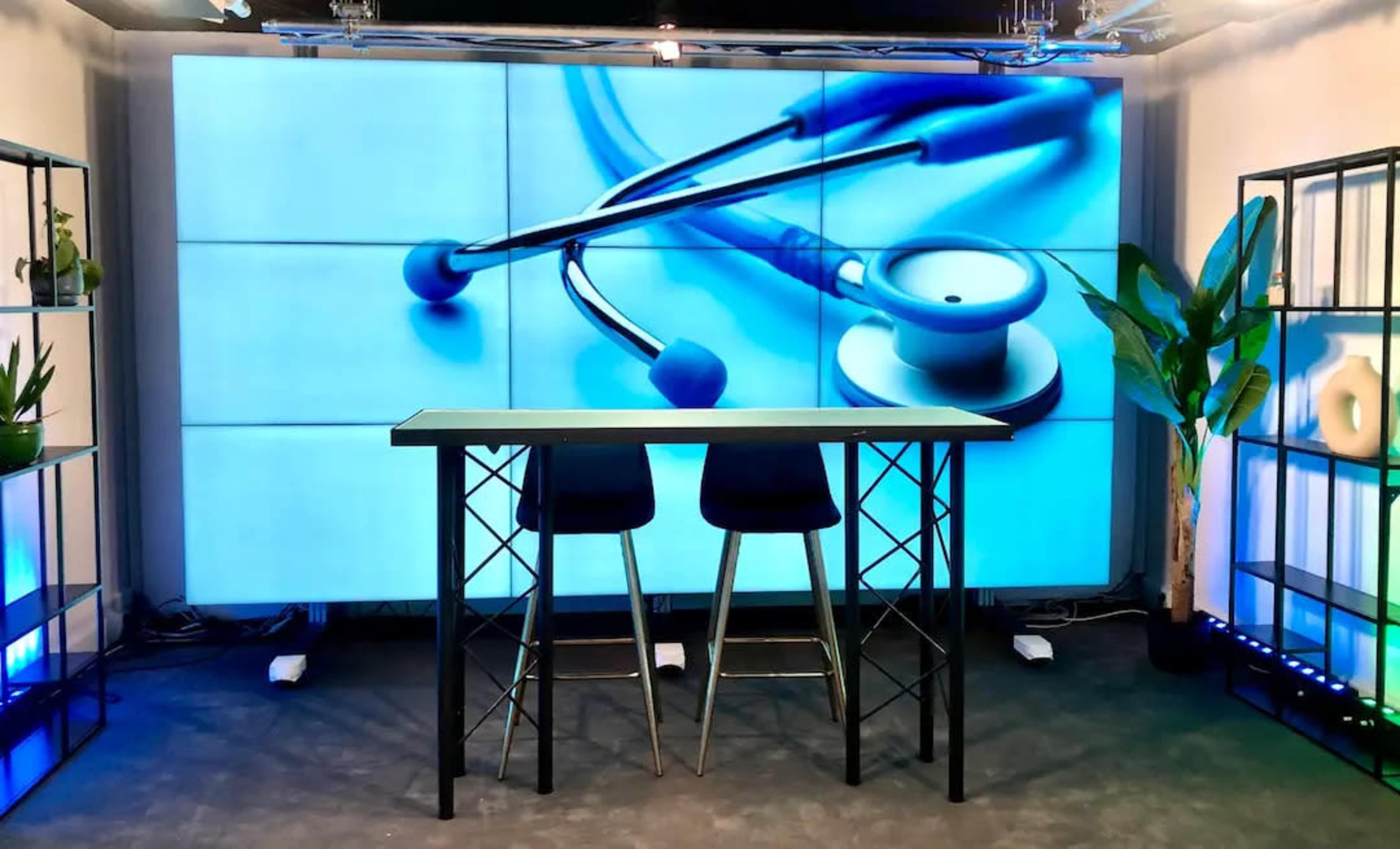A stage setting with a large screen displaying a stethoscope, two high stools, and green plants in black shelving units.