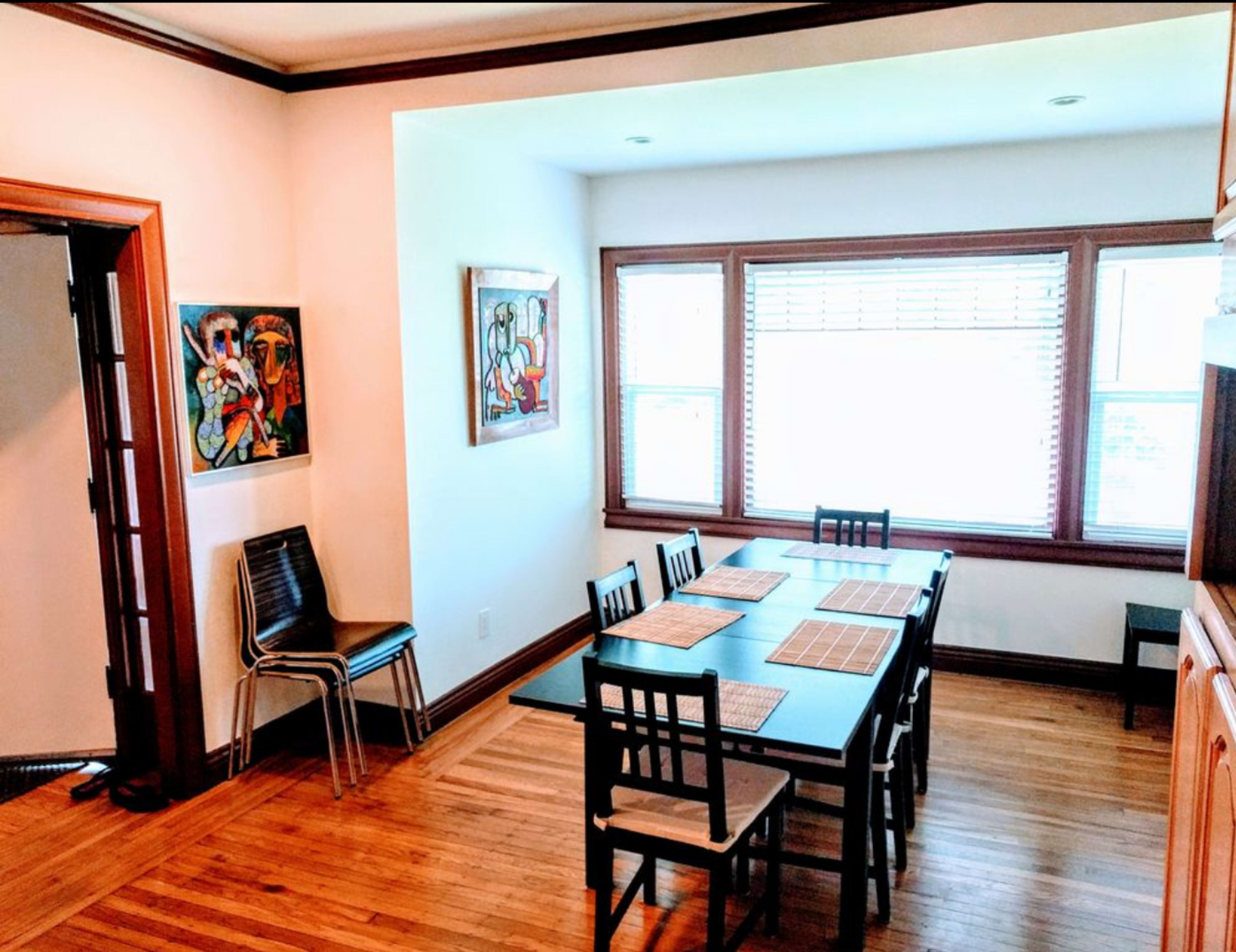 A dining area featuring a rectangular table with several chairs, a window with wooden blinds, and artwork on the walls.