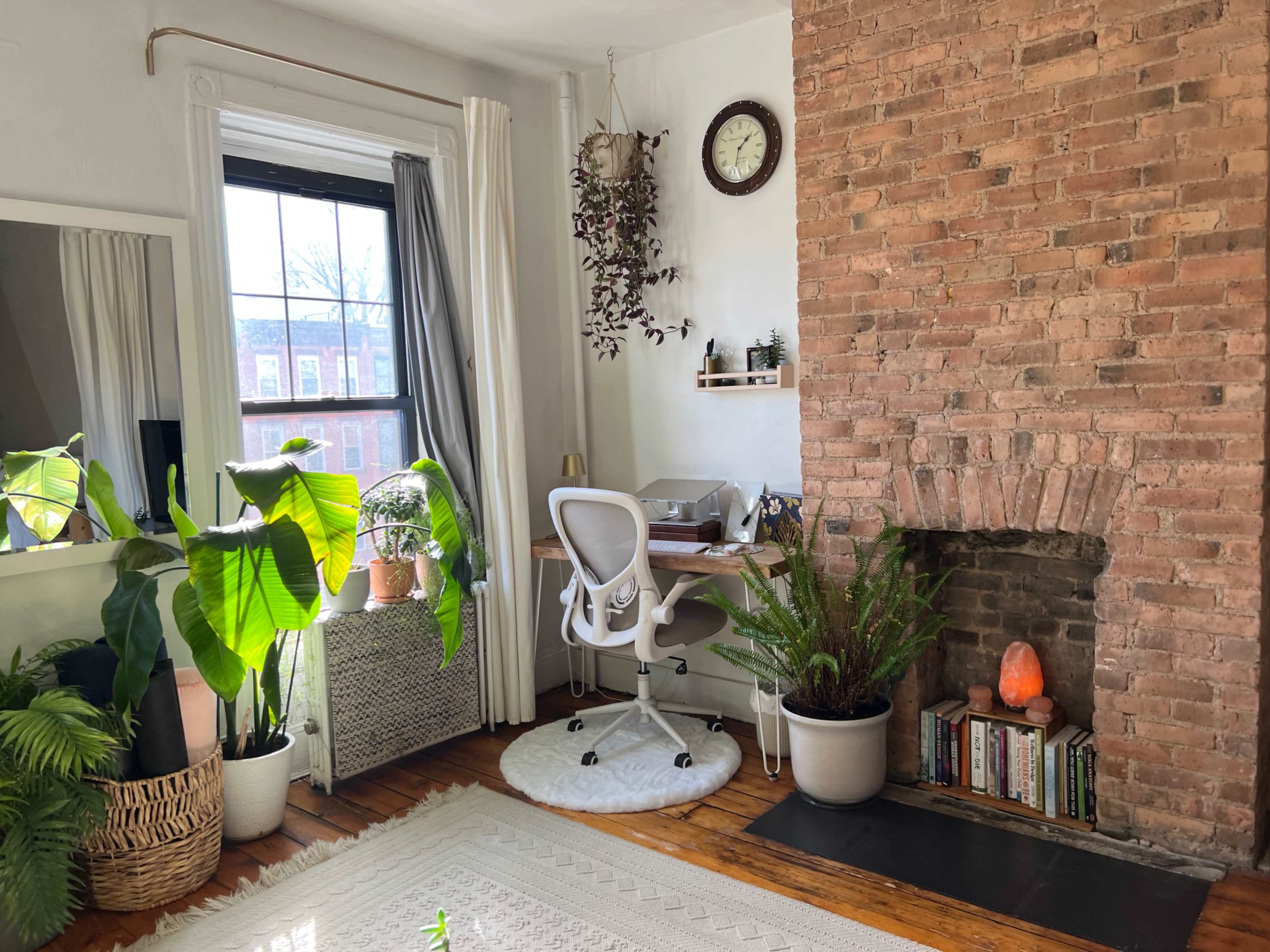 A home office features a desk with a chair next to a brick wall, adorned with plants and a decorative clock.