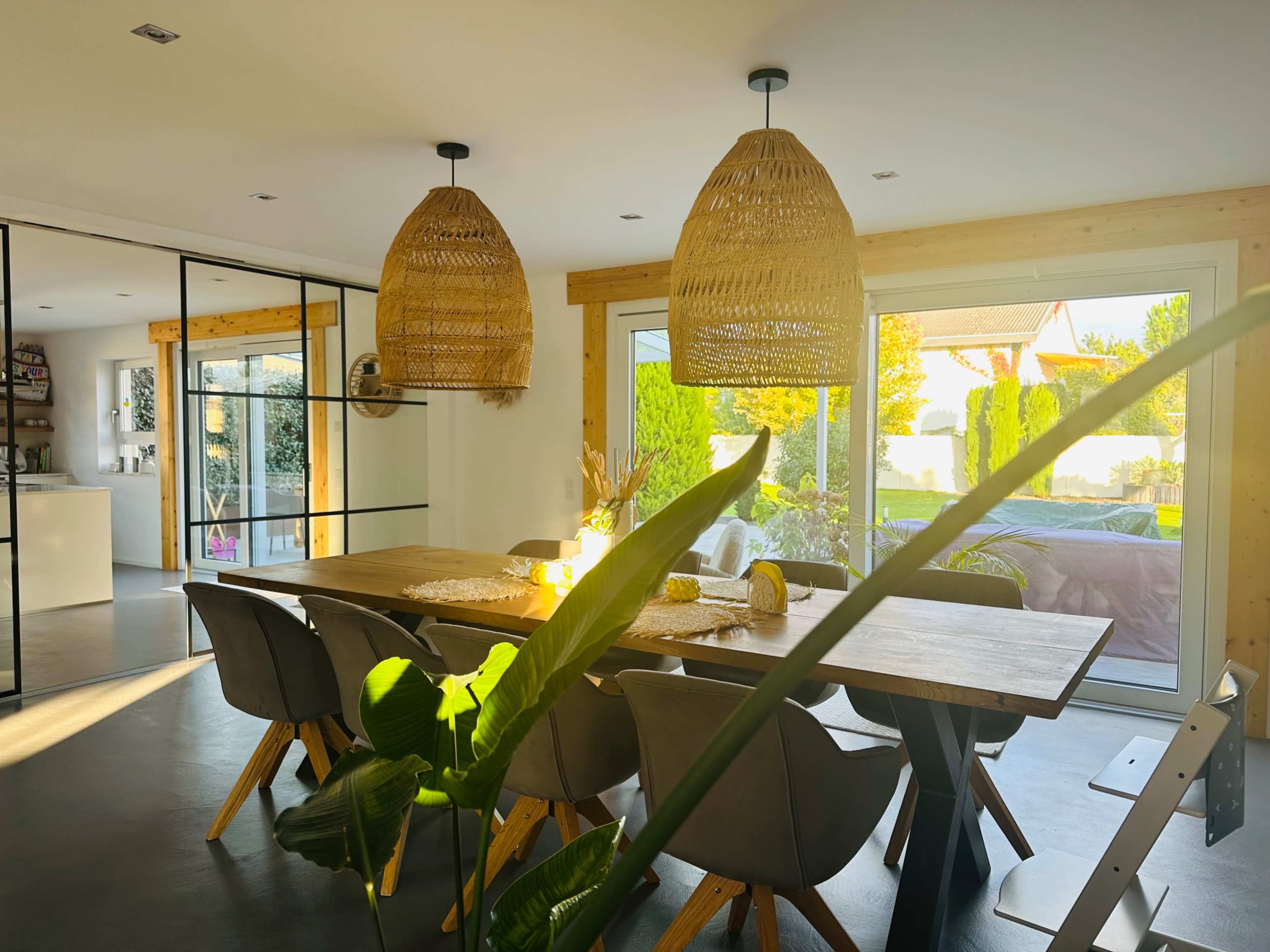 A modern dining area features a large wooden table surrounded by upholstered chairs, with two large woven pendant lights above and sliding glass doors leading to an outdoor space.