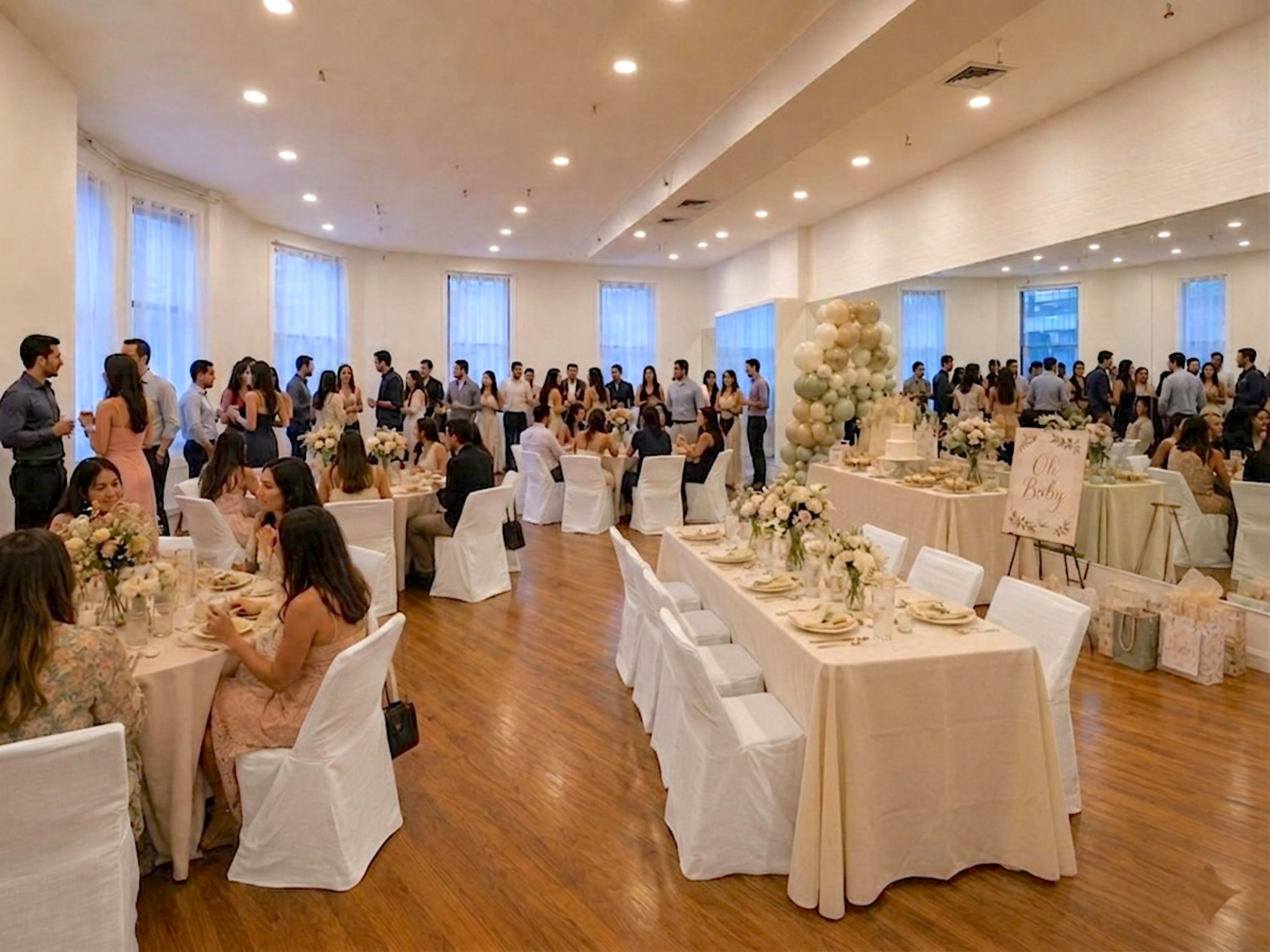 A large, well-lit event hall is filled with guests seated at elegantly set tables, while others mingle near decorated displays and balloons.