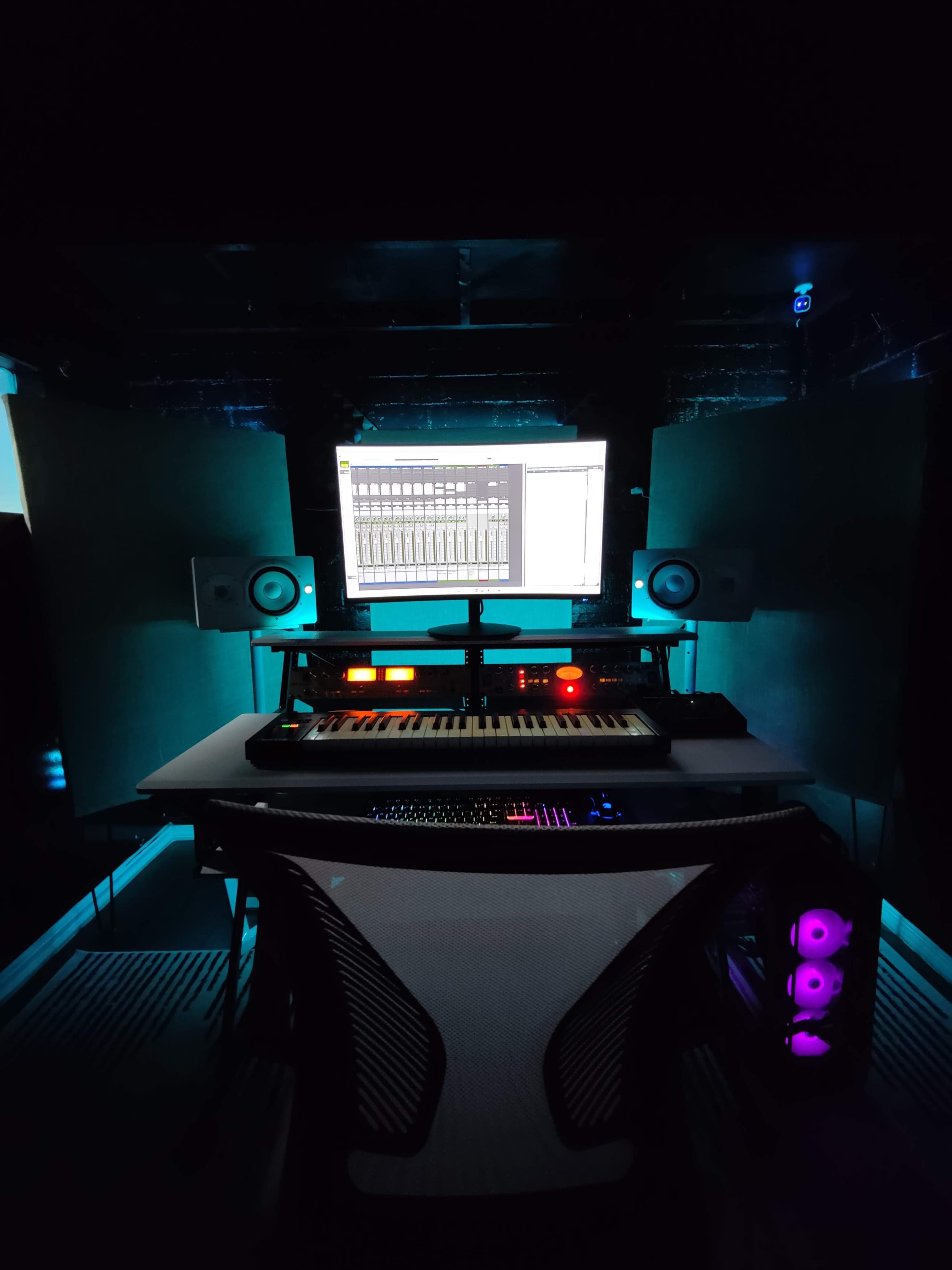 A music production studio features a computer monitor, a MIDI keyboard, and speaker monitors against a dark backdrop.
