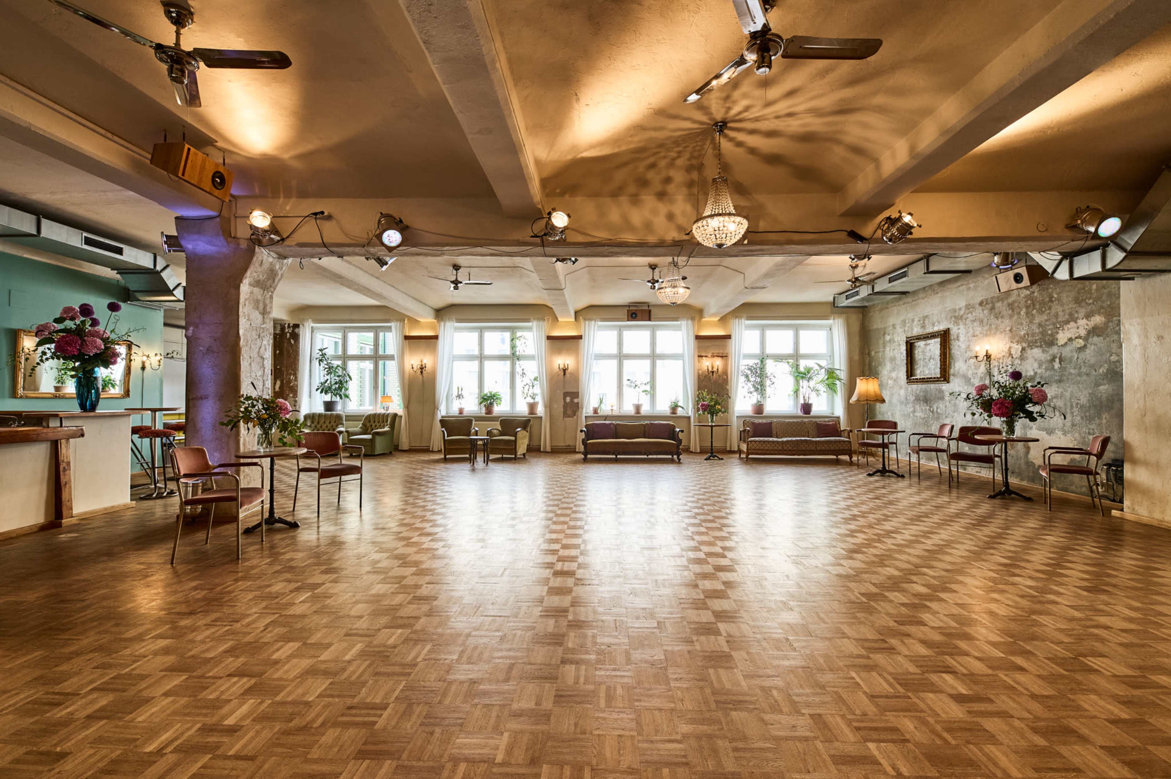 An empty dance hall with wooden flooring, large windows, and seating along the walls, adorned with flowers.