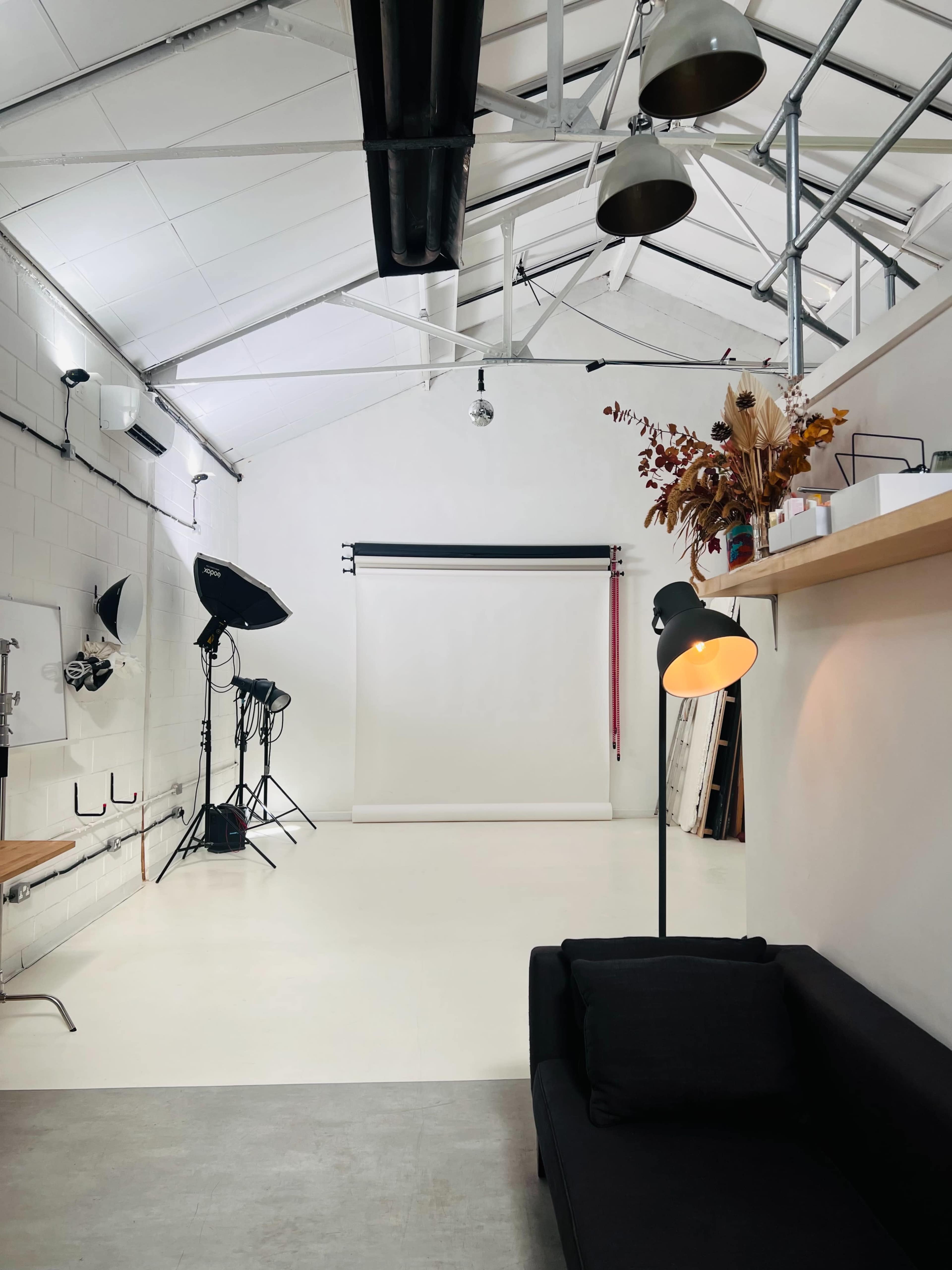 The image shows a photography studio with a white backdrop, lighting equipment, and a black sofa against one wall.