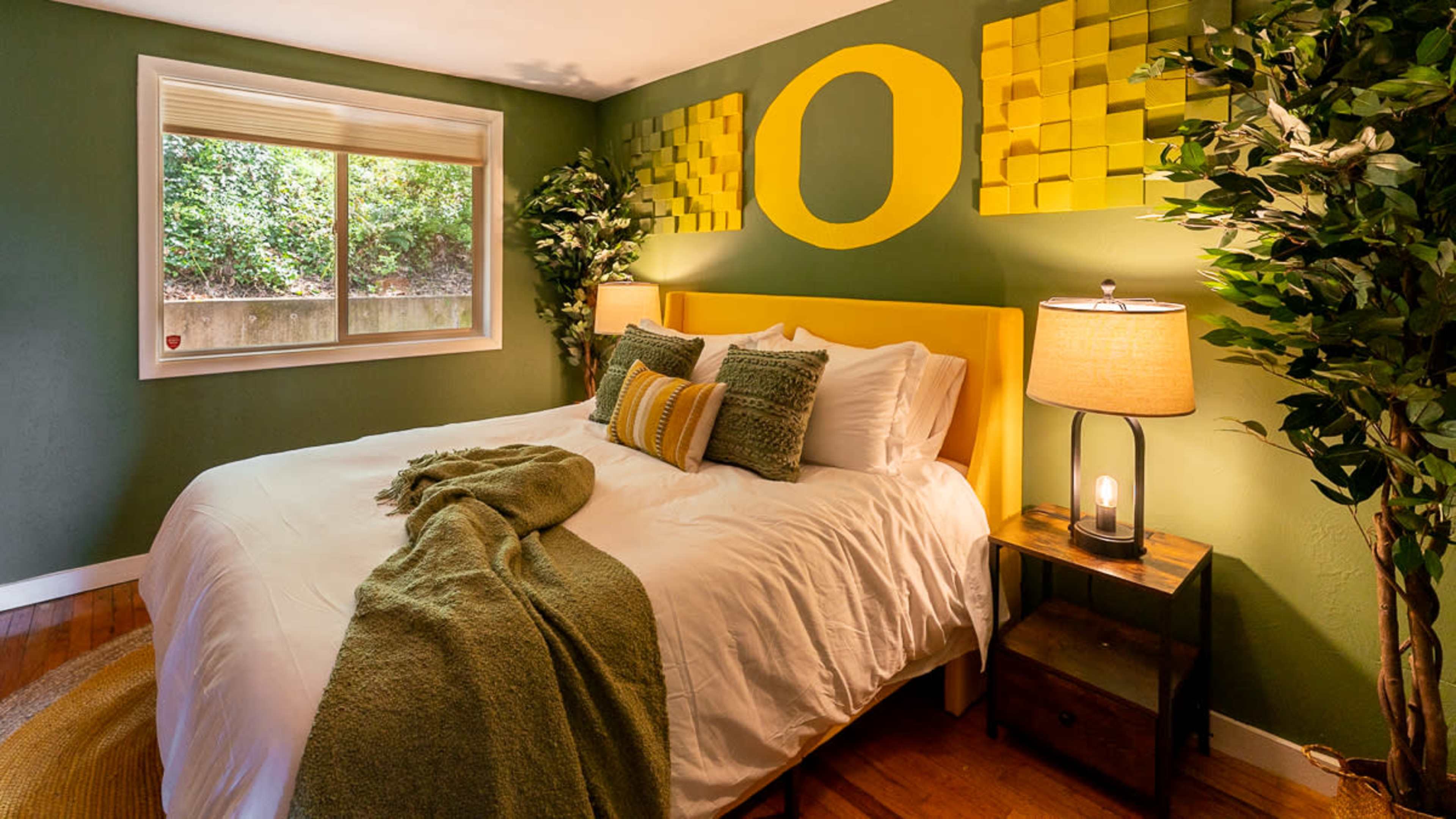 A bedroom with a yellow bed frame, green walls, decorative square patterns, and a window overlooking greenery.