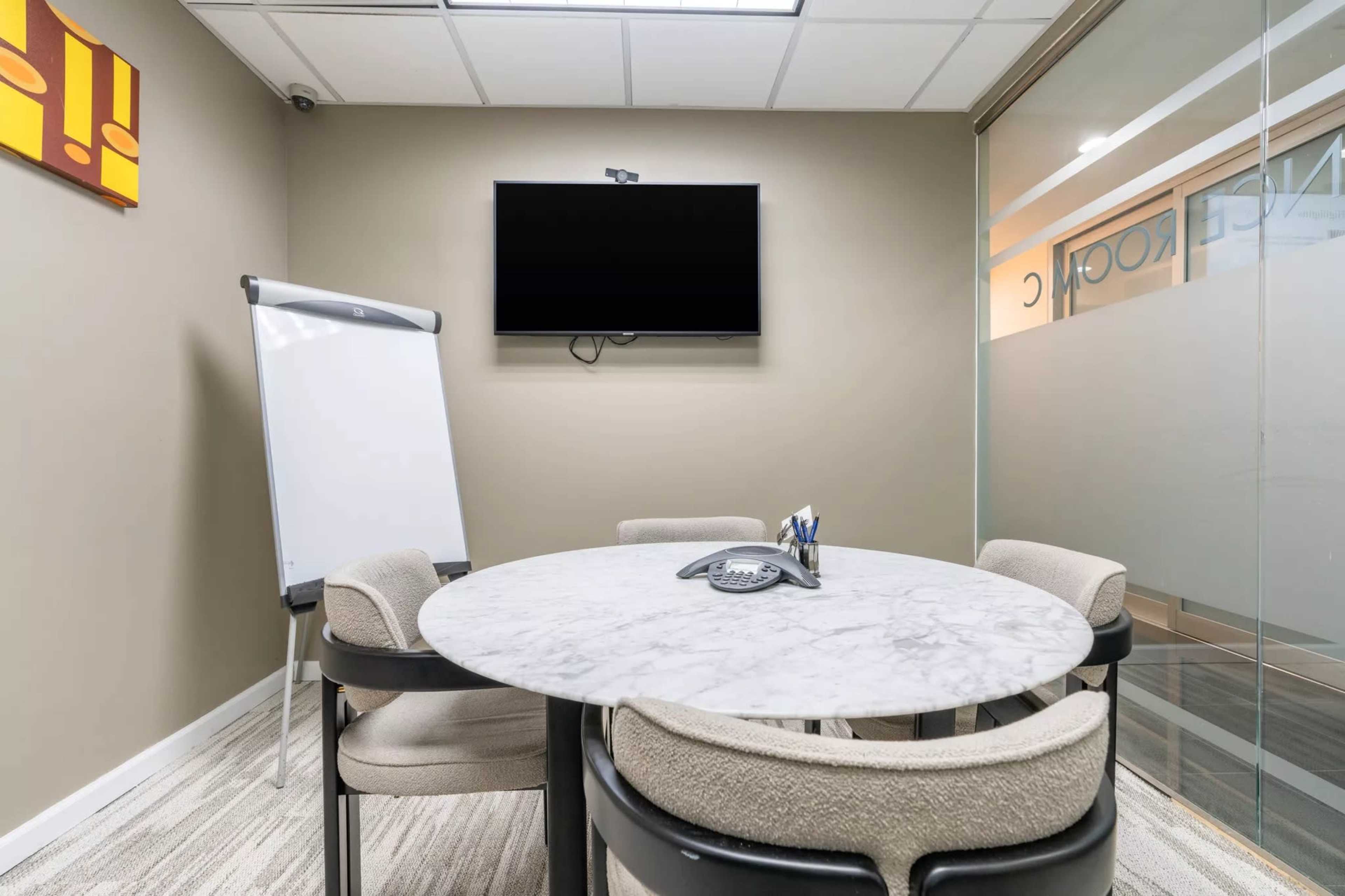 The image shows a small conference room featuring a round marble table, four upholstered chairs, a whiteboard, and a wall-mounted television.