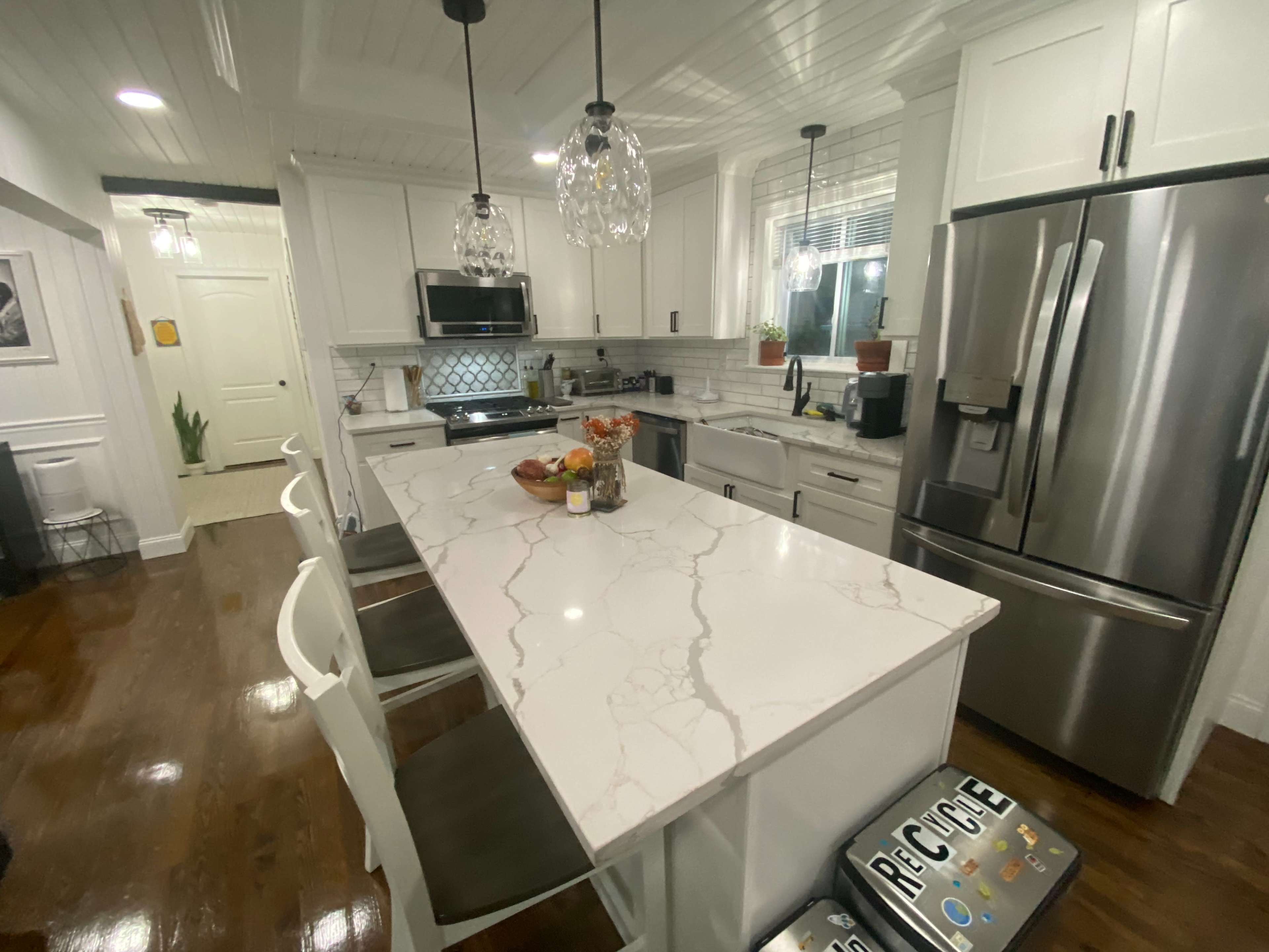 A modern kitchen features white cabinetry, a large marble island with seating, stainless steel appliances, and pendant lighting.