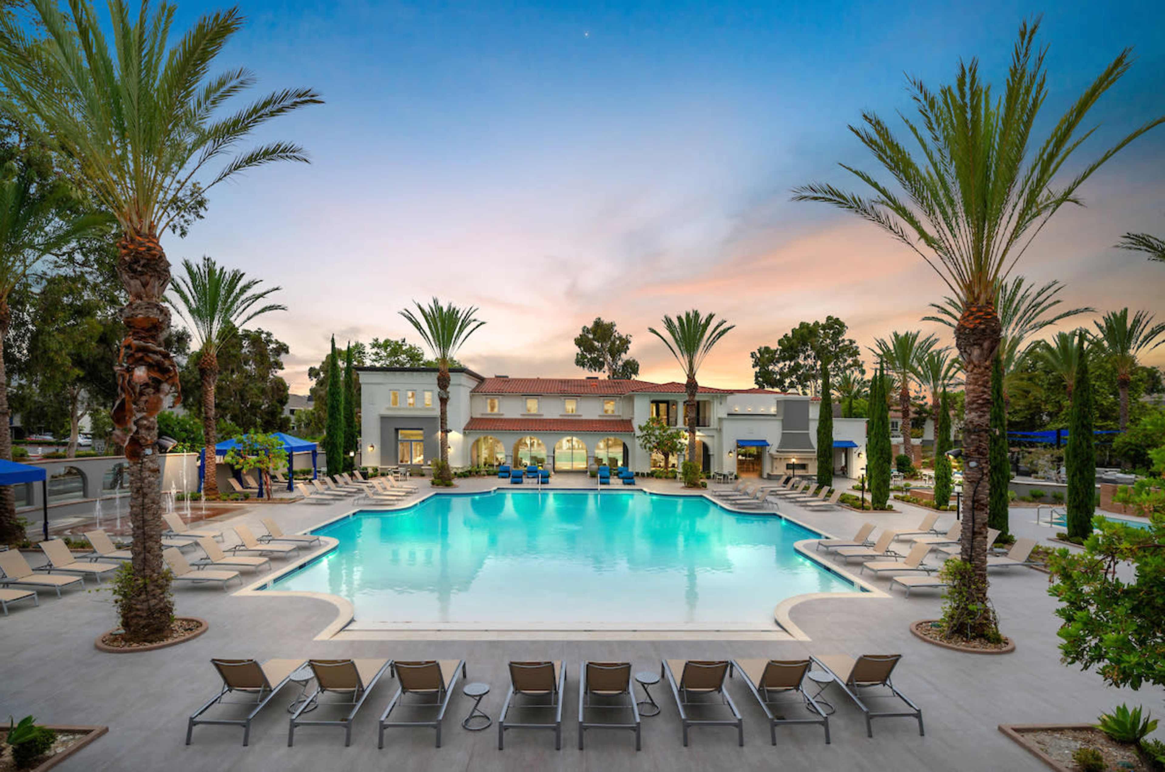 A well-lit swimming pool surrounded by palm trees and lounge chairs, with a modern building in the background under a sunset sky.