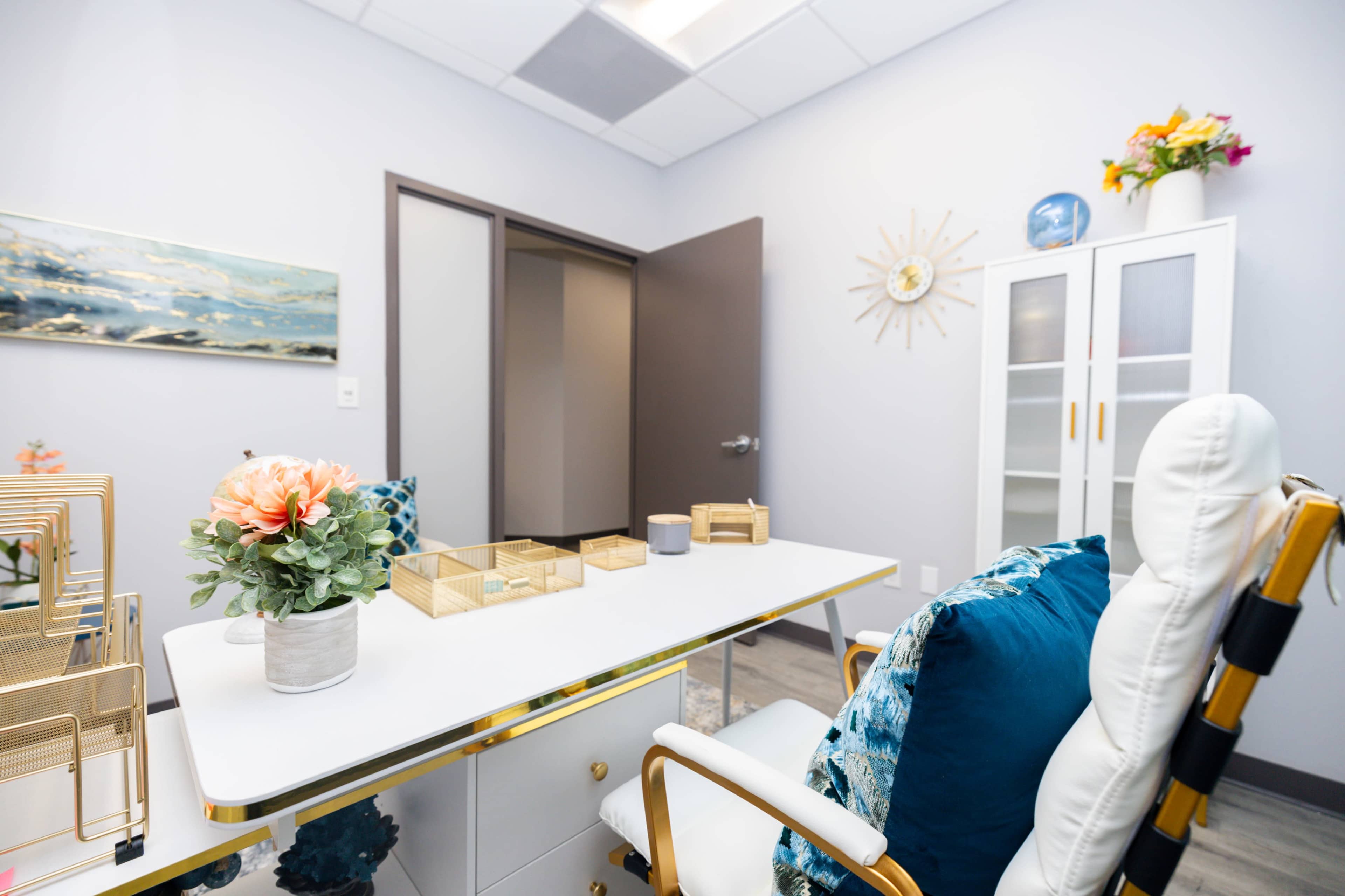 The image shows a modern office interior with a white desk, a chair, decorative plants, and a light-colored wall adorned with wall art and a cabinet.