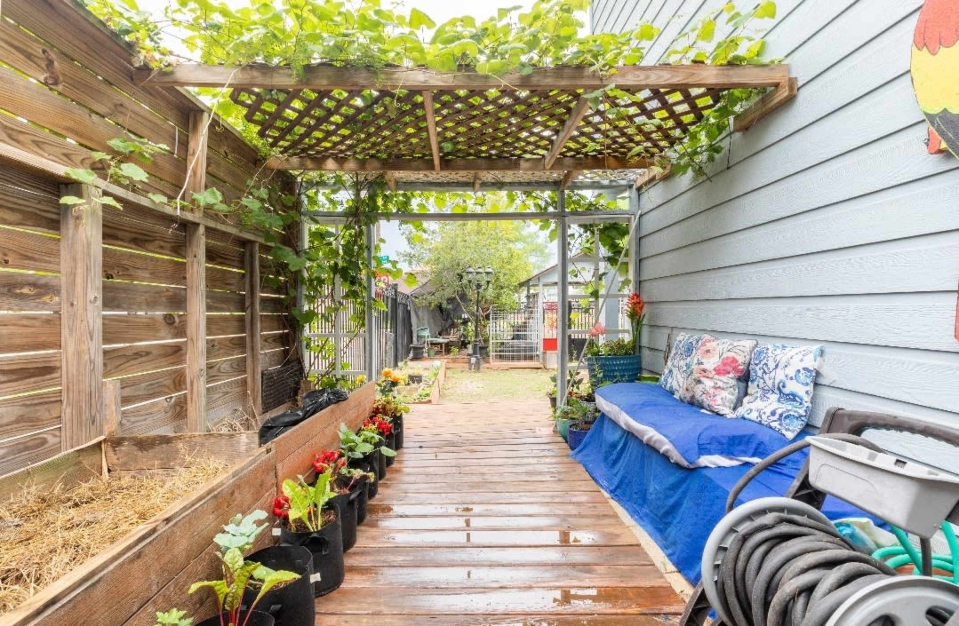 The image shows a wooden pathway leading through a garden area with potted plants on either side and a trellis overhead covered in greenery.
