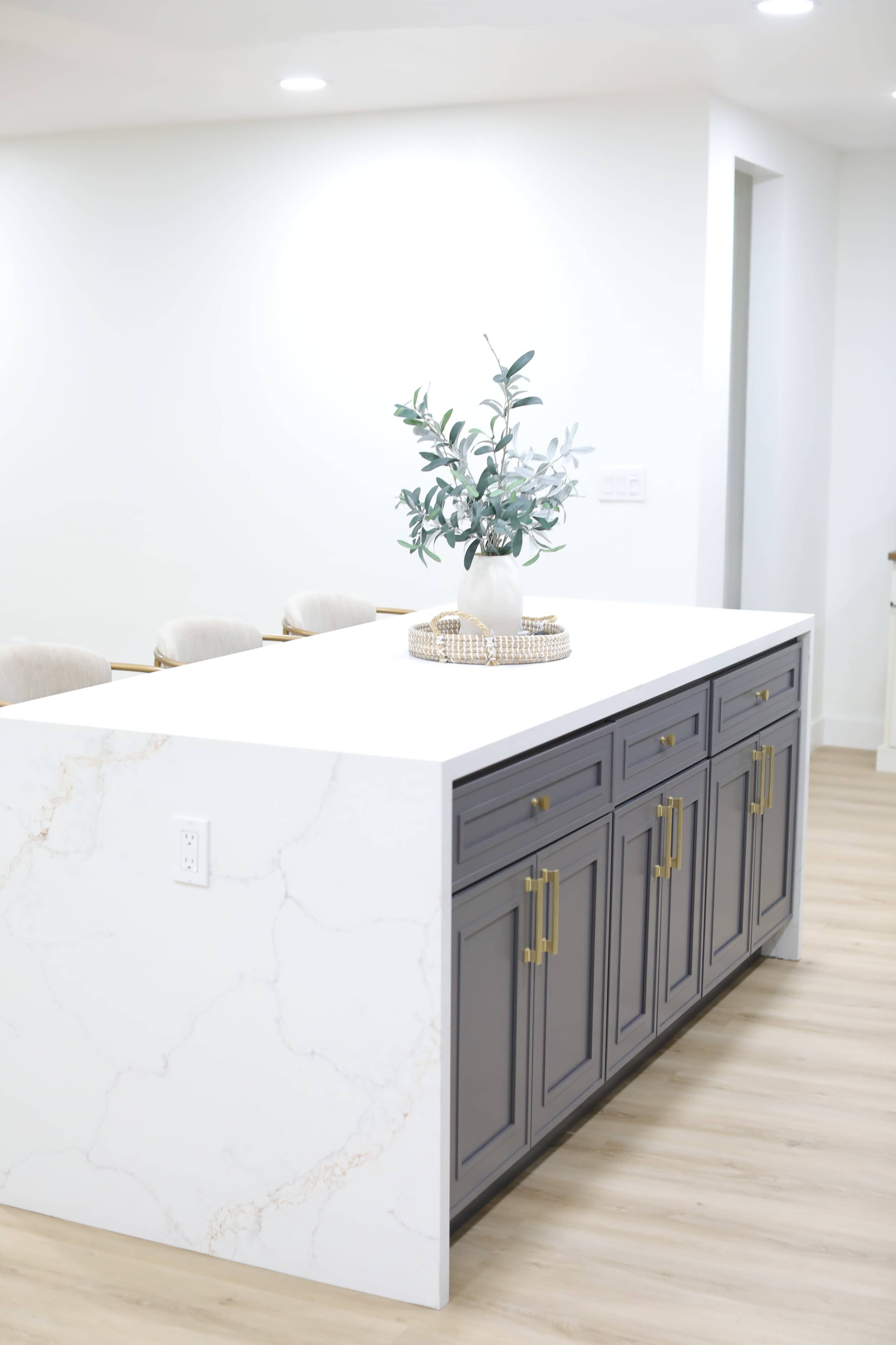 The image shows a modern kitchen with a large white marble-topped island featuring gray cabinets and a decorative vase of greenery on top.