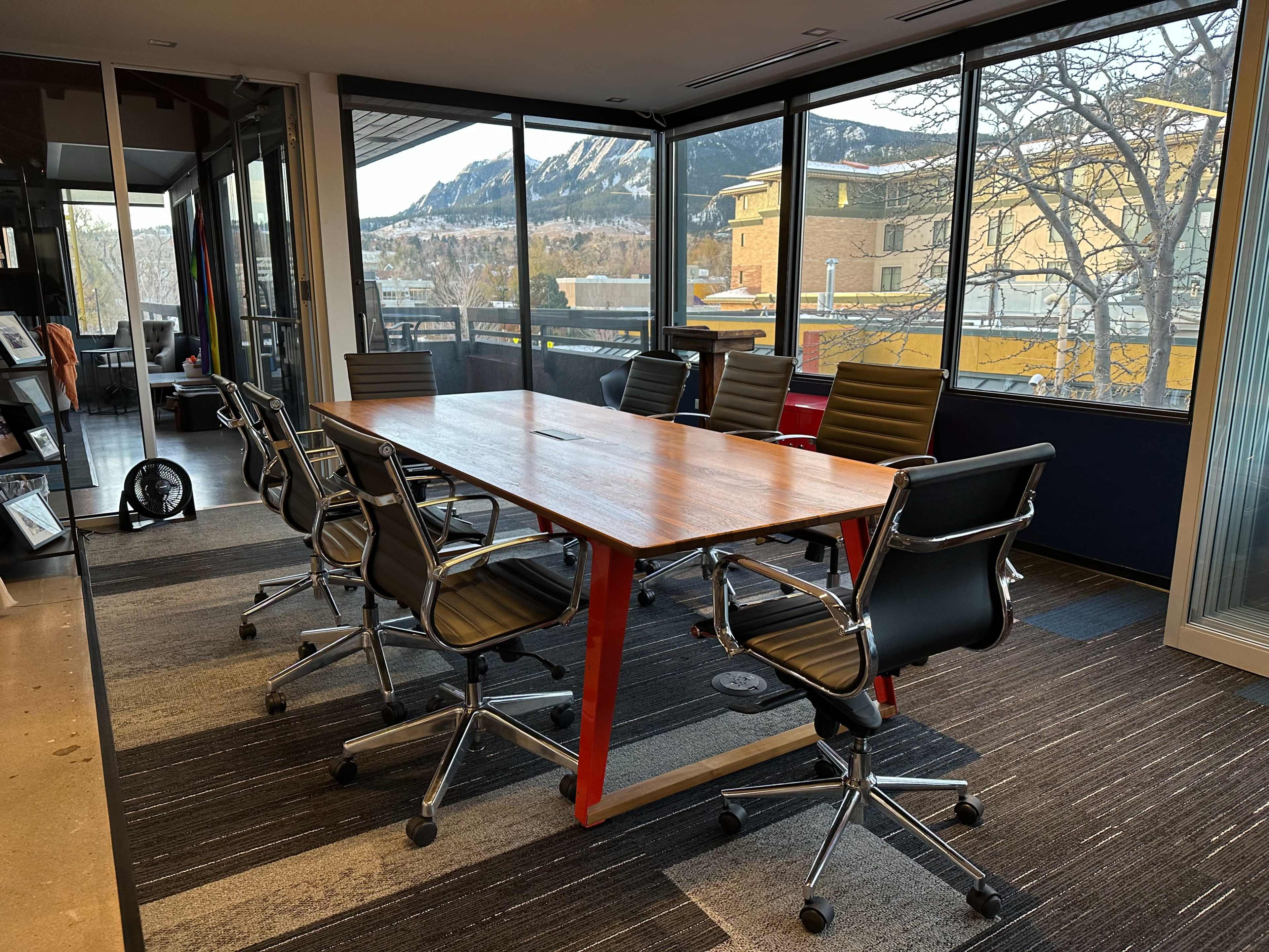 A conference room features a wooden table surrounded by six black ergonomic chairs, with large windows displaying mountains in the background.
