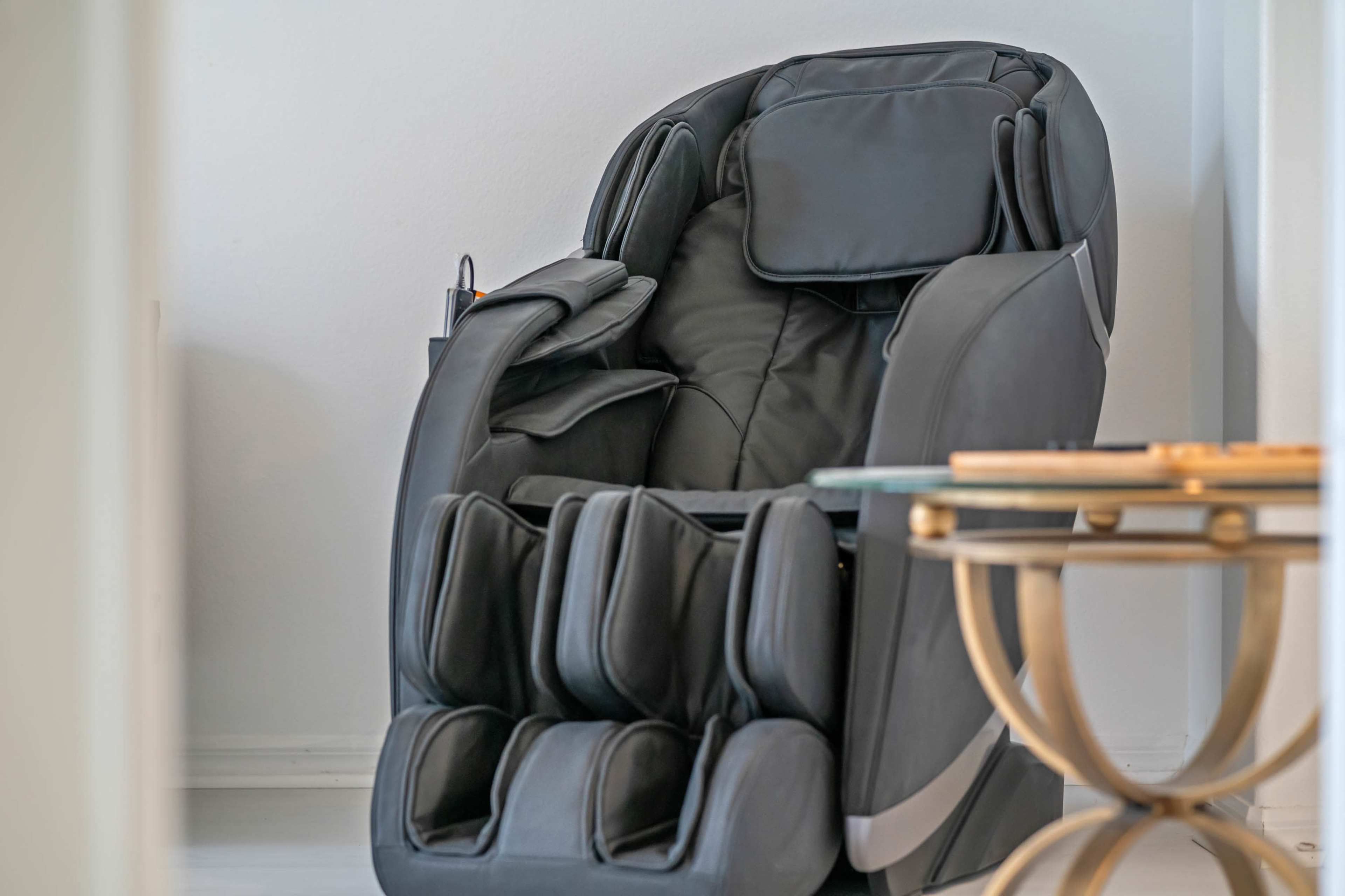 A black massage chair is positioned next to a small, round side table in a light-colored room.