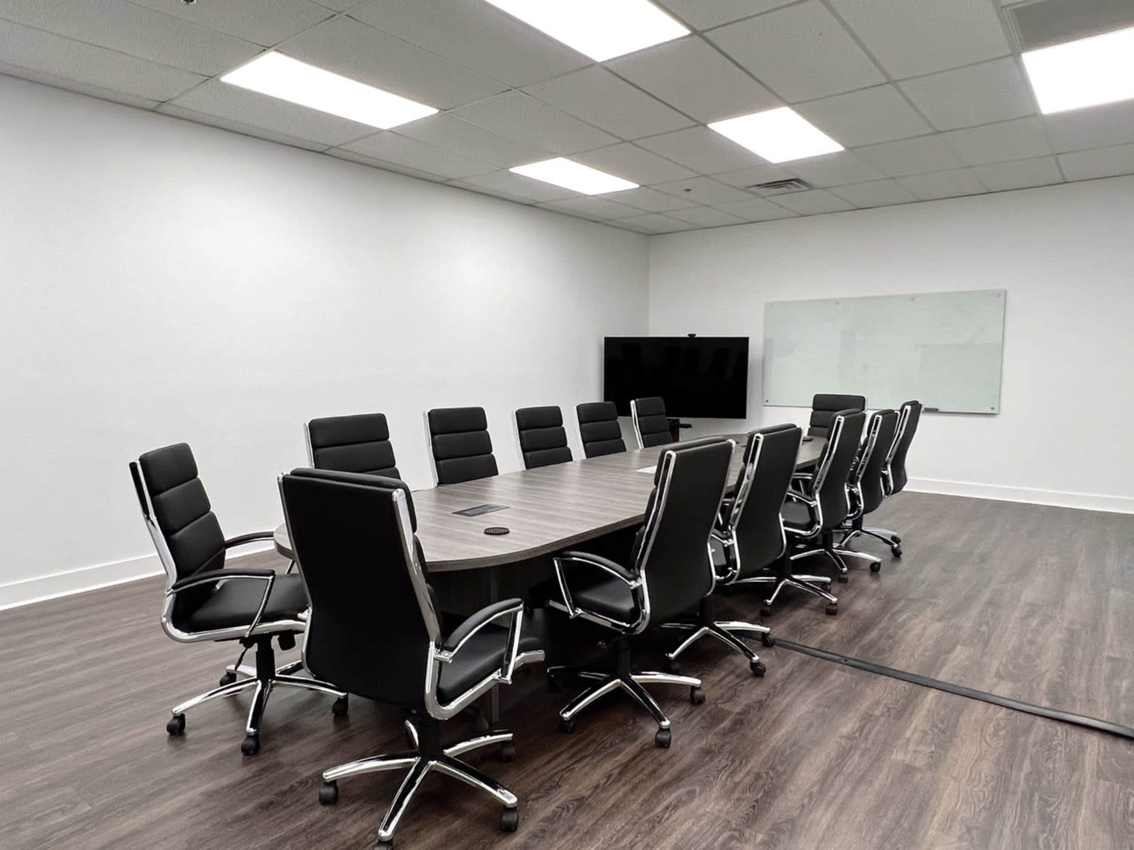 A spacious, modern conference room with a large oval table surrounded by twelve black leather chairs, a wall-mounted screen, and a whiteboard.