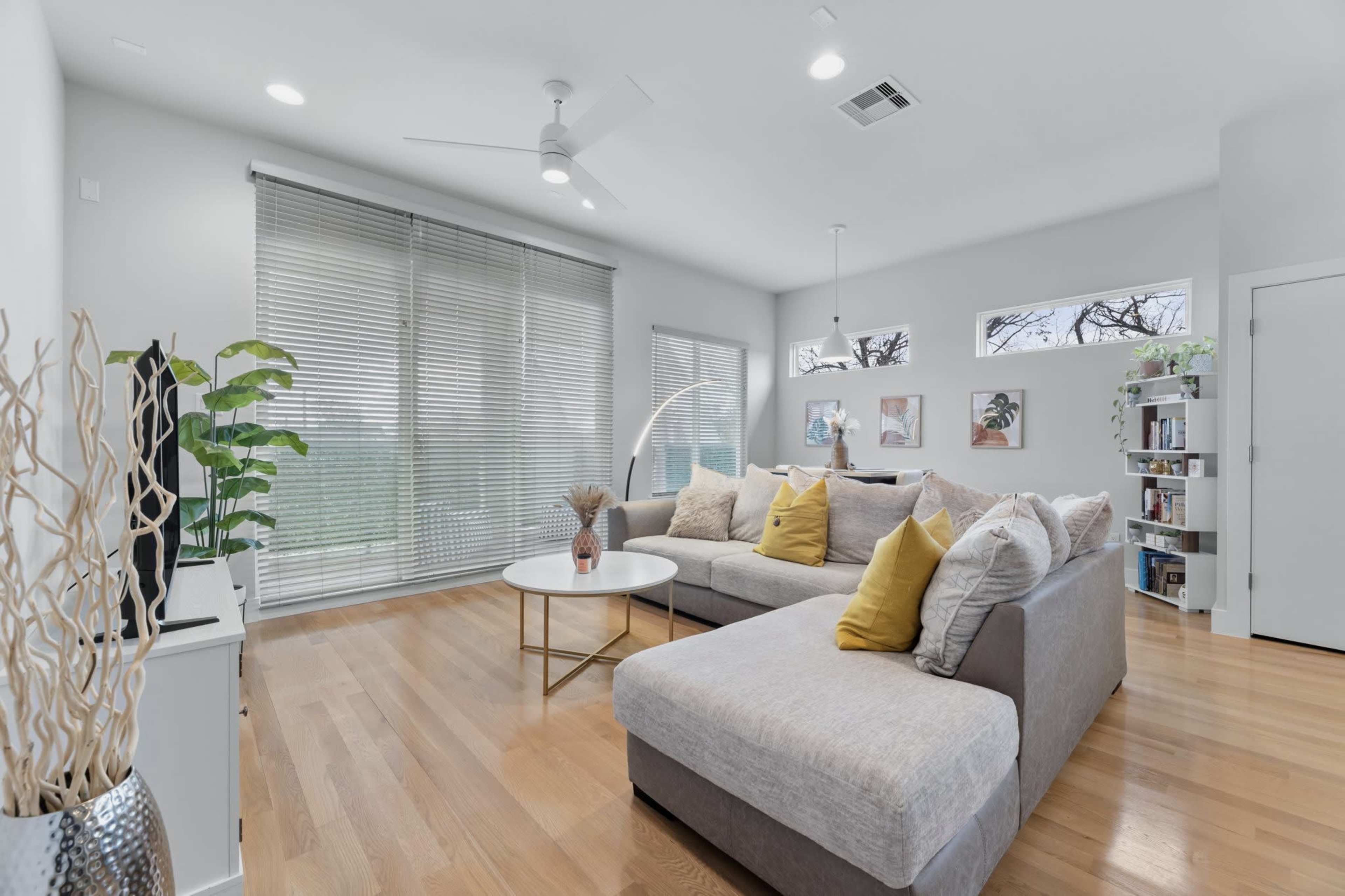 A modern living room features a large sectional sofa with yellow accent pillows, a round coffee table, and shelves displaying books and decorative items, complemented by large windows with blinds.