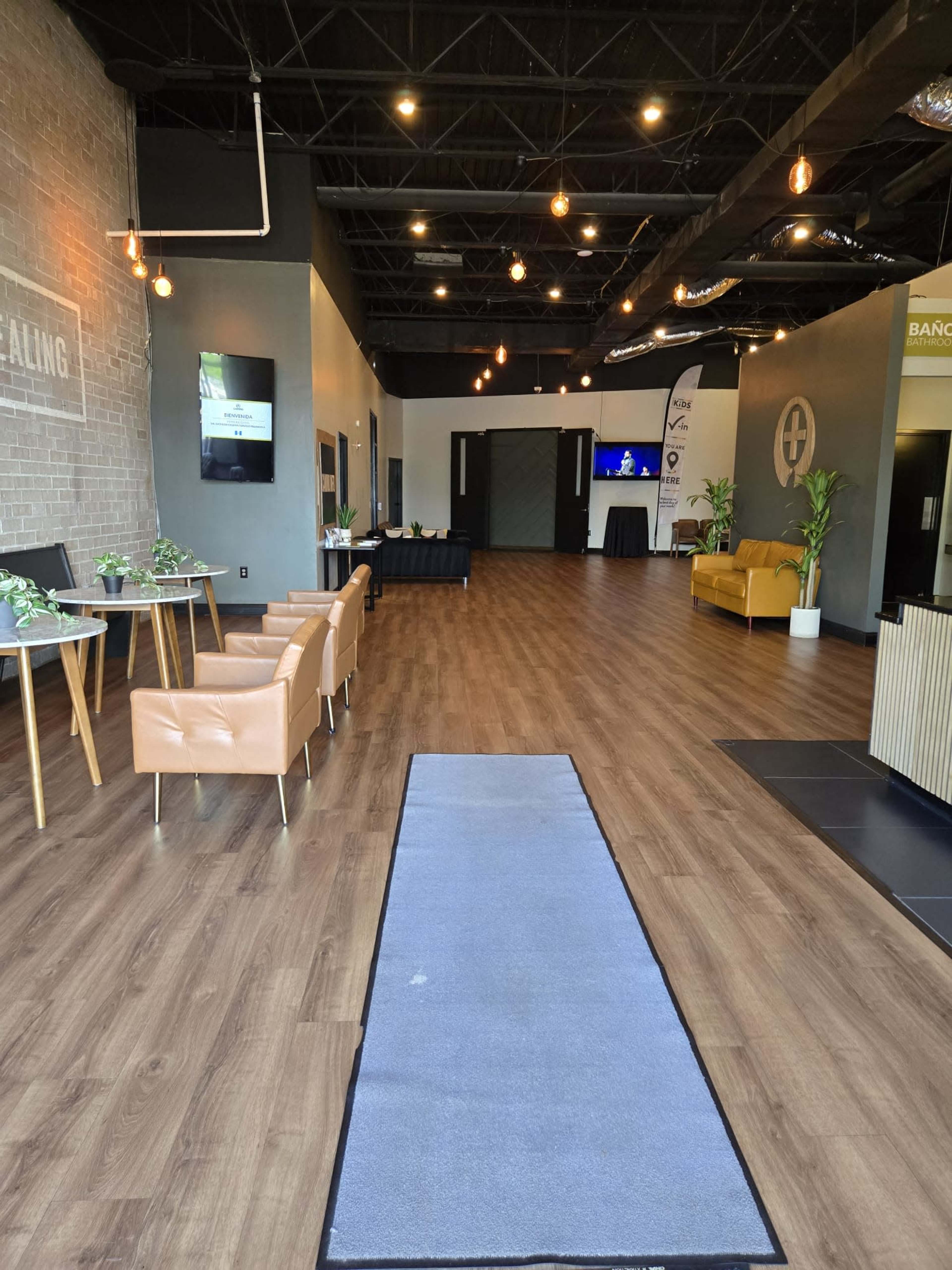 The image shows a modern interior space with wooden flooring, a long gray runner rug, several seating areas with brown chairs, and a reception area at the back.