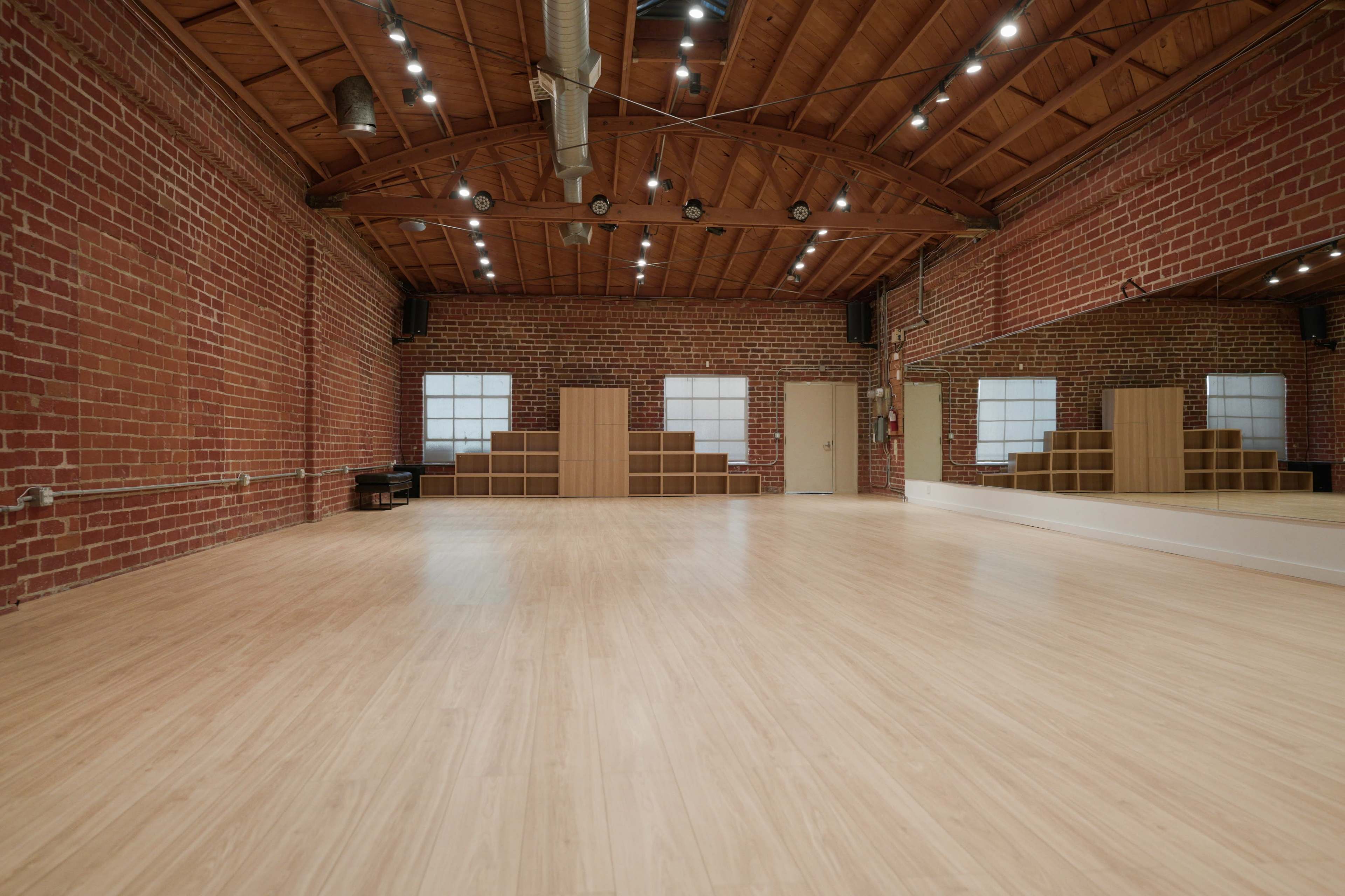 A spacious dance studio with wooden floors, exposed brick walls, and multiple bright lights overhead.