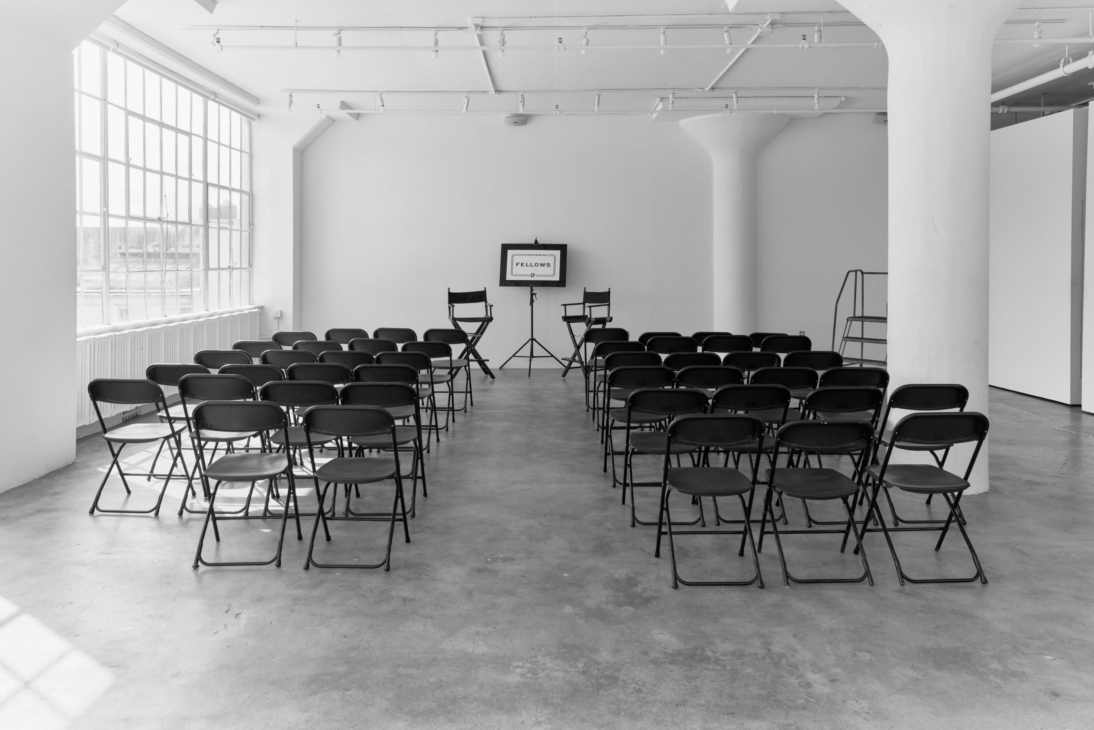 A minimalist room with a row of black folding chairs facing a projector screen on a stand.