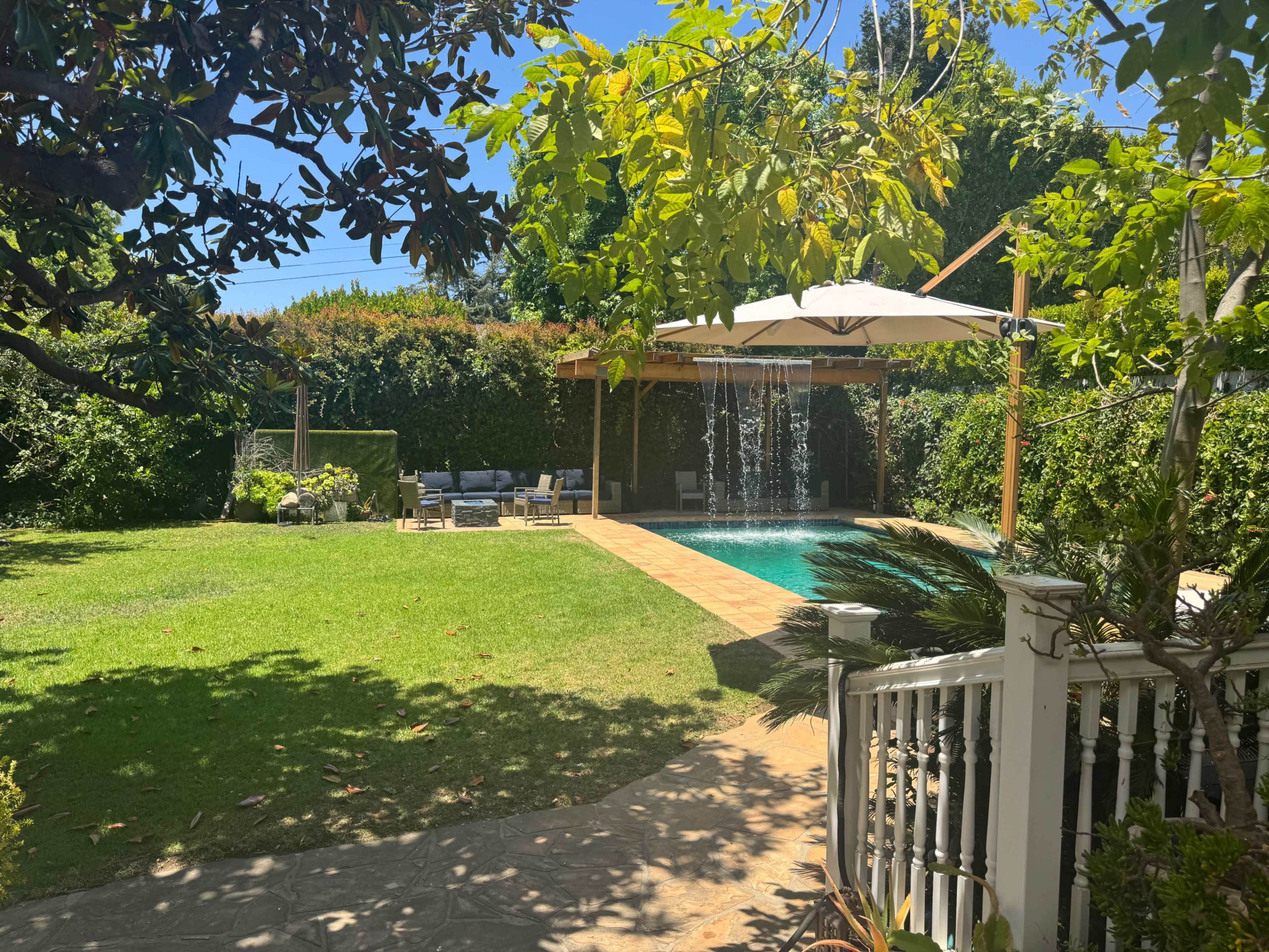 The image shows a residential backyard with a swimming pool, patio furniture, and a waterfall feature surrounded by lush greenery.