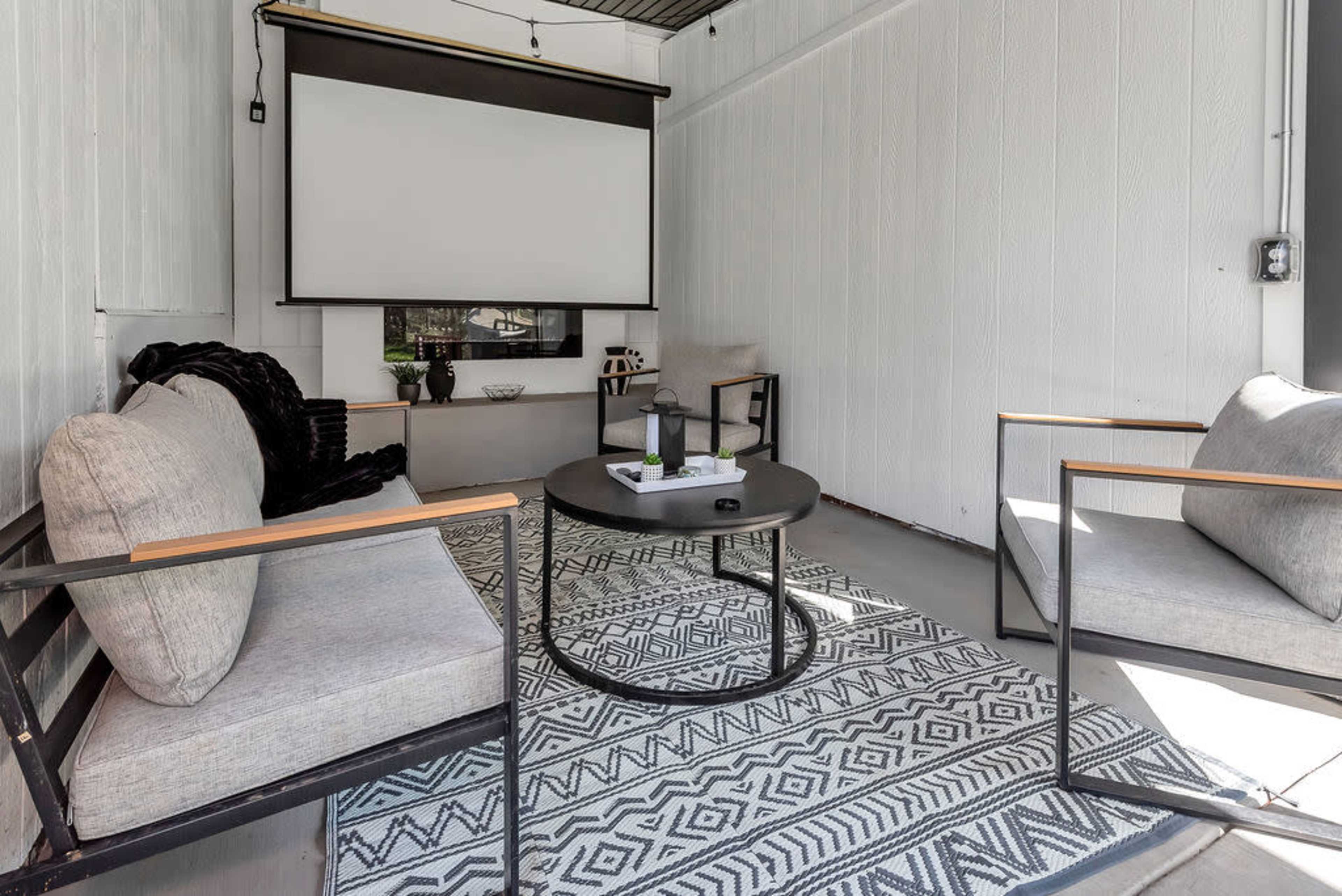 A modern outdoor seating area with two chairs and a round coffee table, featuring a large retractable screen mounted on the wall.