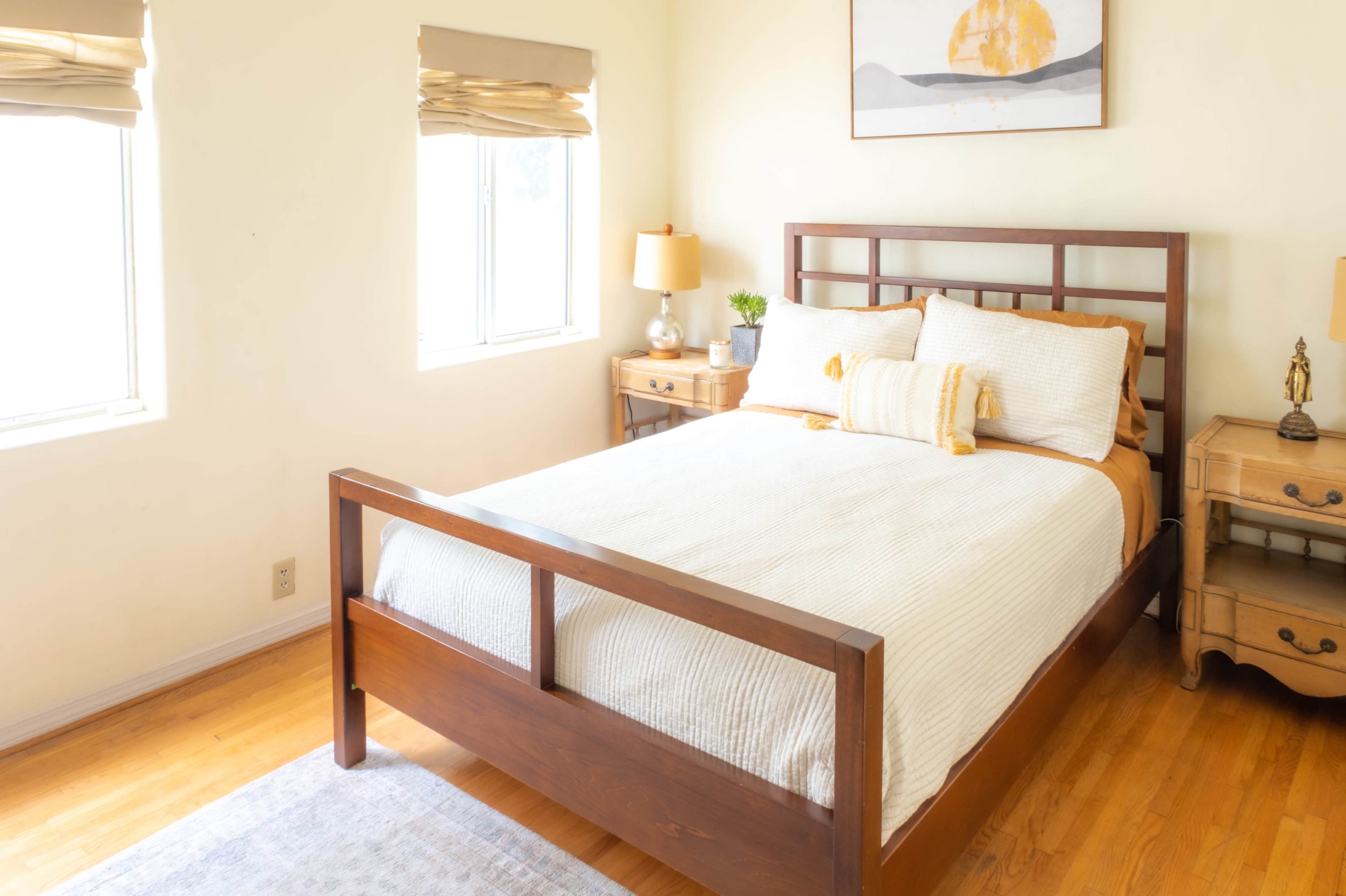 A neatly arranged bedroom features a wooden bed with white bedding, two bedside lamps, and a small table beside a window.