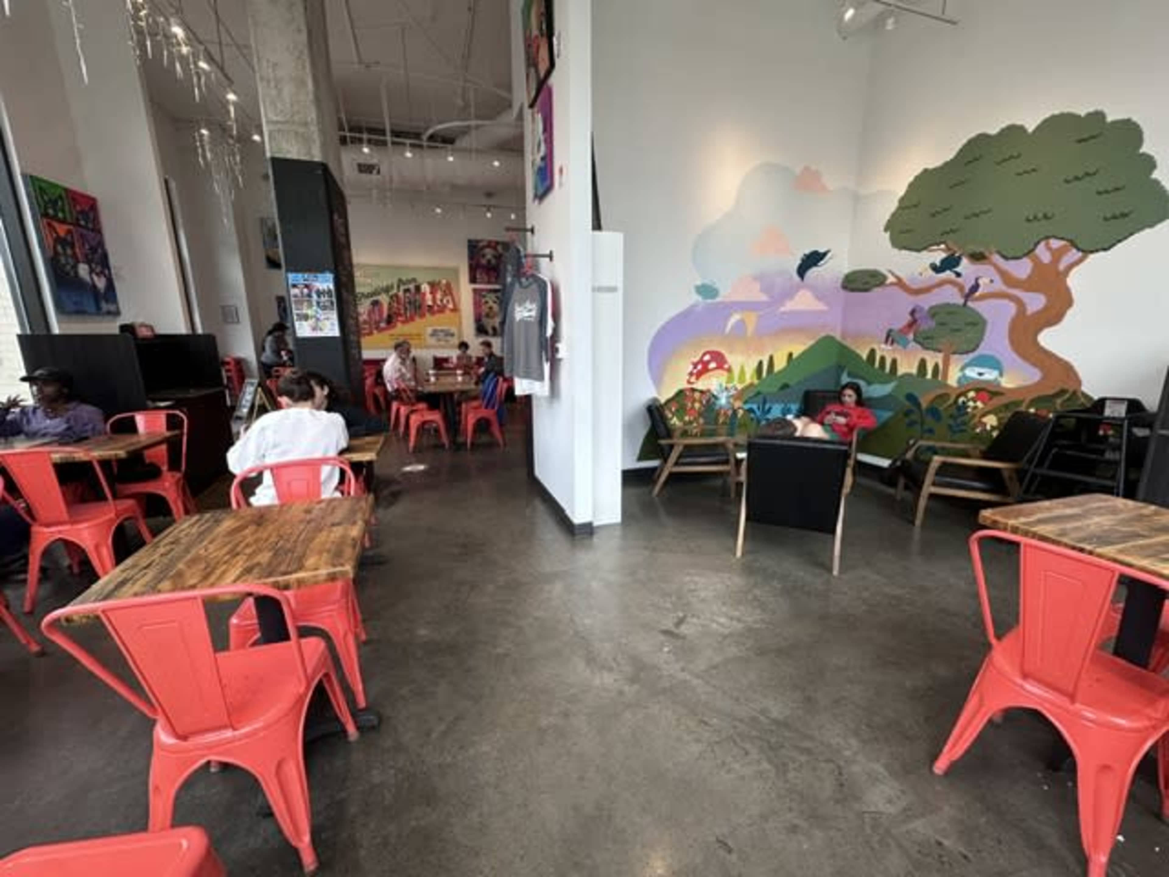 The interior of a café features red metal chairs, wooden tables, and a mural depicting a colorful landscape on one wall, with several patrons seated at various tables.