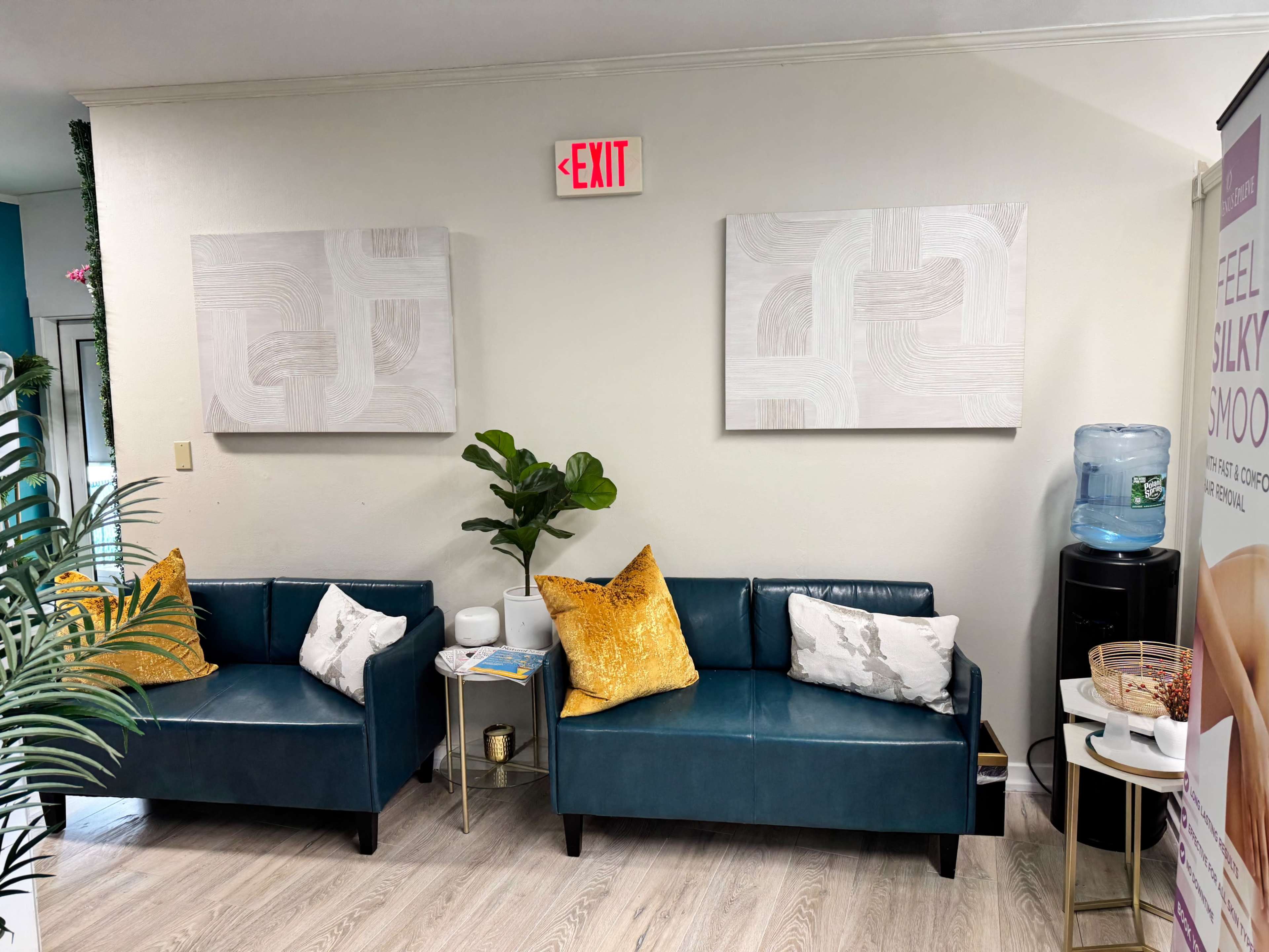 The image shows a waiting area with two teal couches, decorative pillows, a potted plant, and a water cooler against a light-colored wall adorned with two abstract paintings.